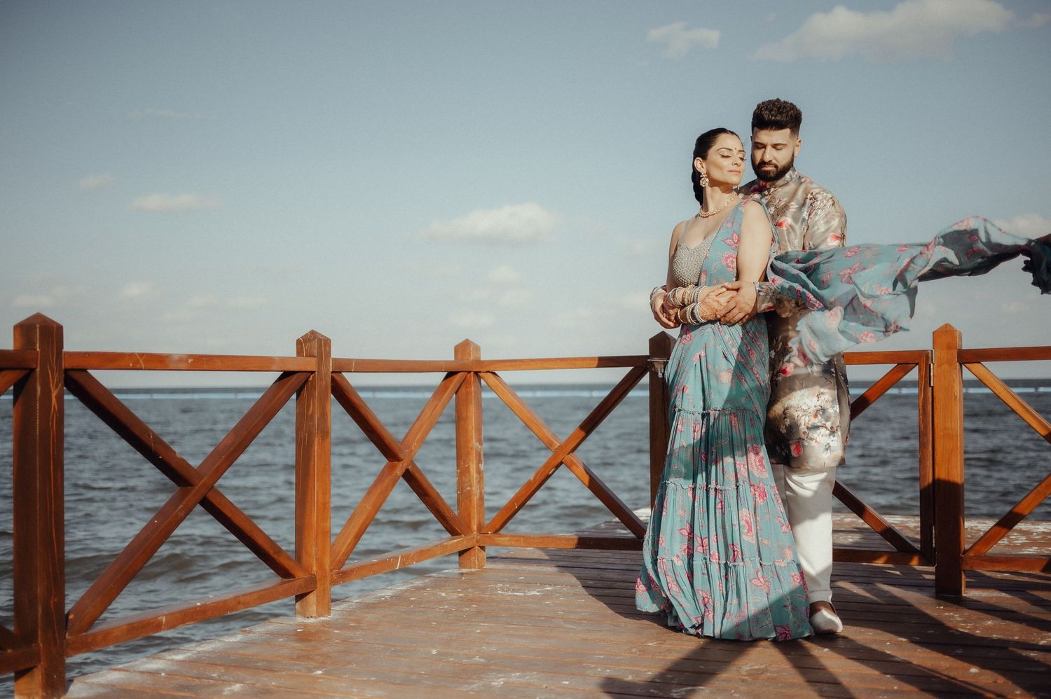 A couple in relaxed clothing embraces while standing on a wooden pier overlooking the ocean at sunset.