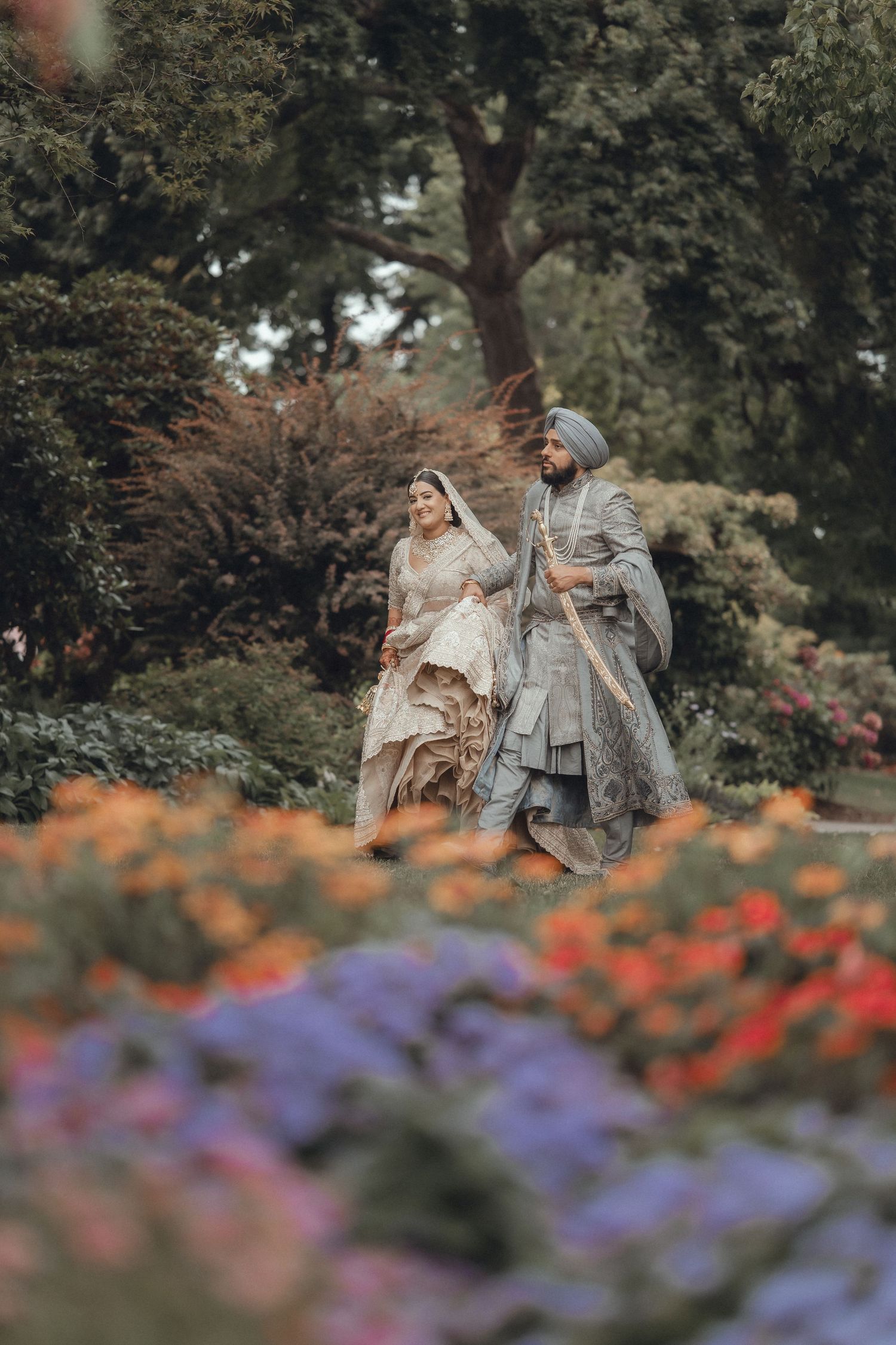 A couple in luxurious traditional attire walking through a colorful garden with purple and orange flowers.