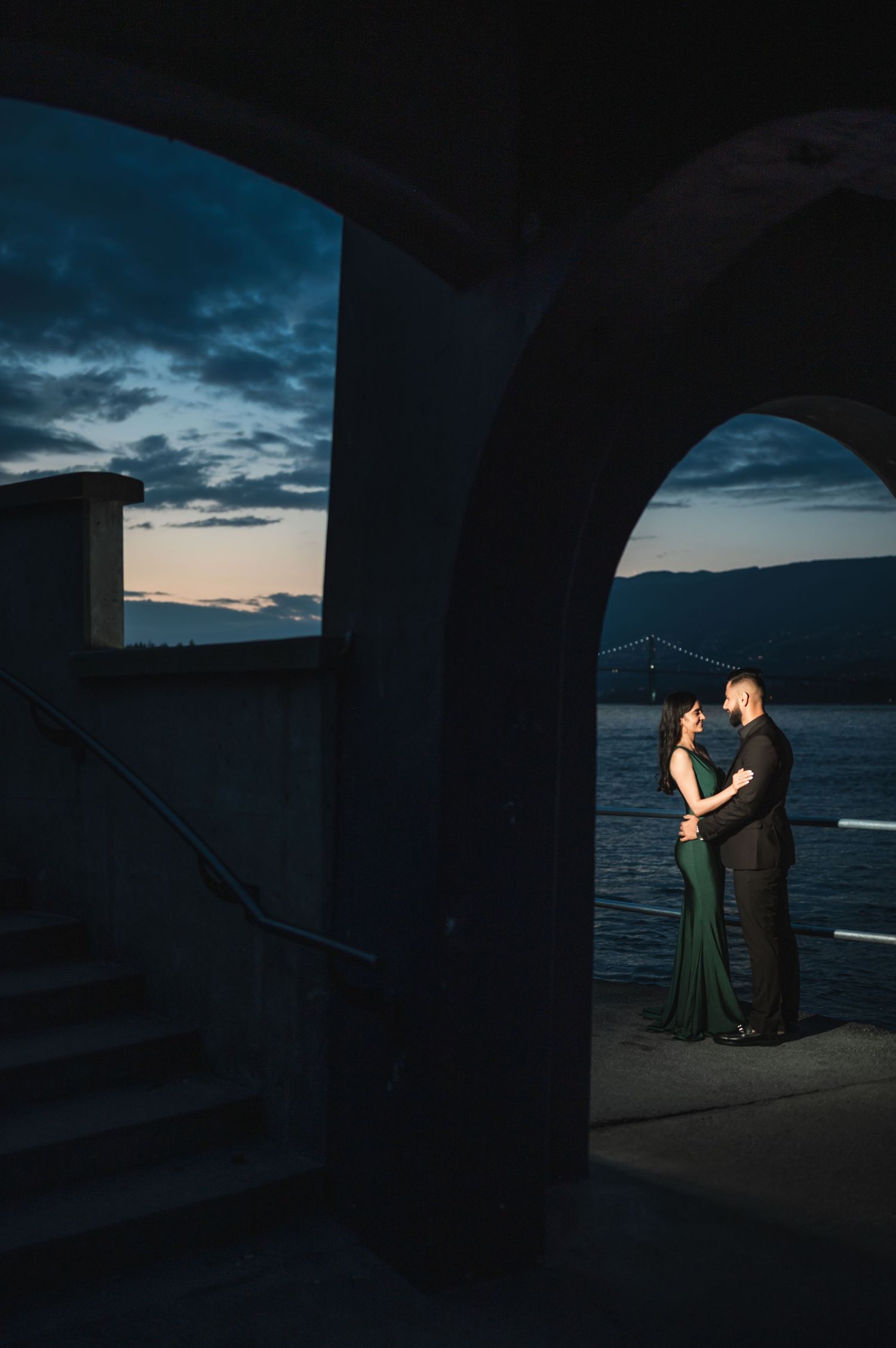 Couple in formal wear embracing under waterfront arches during twilight.