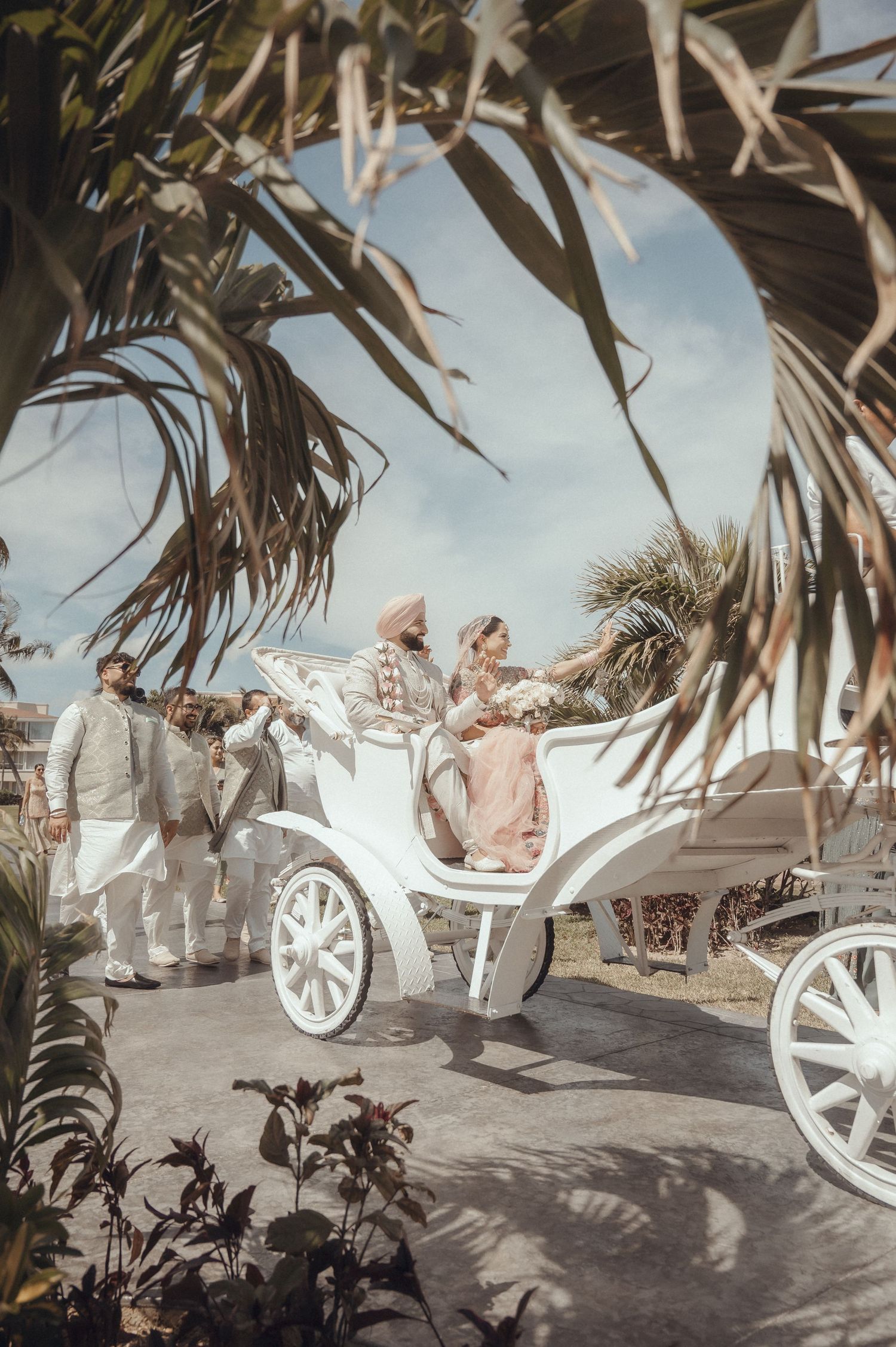 A white horse-drawn carriage stands elegantly among tropical palm trees at a beachfront wedding venue.
