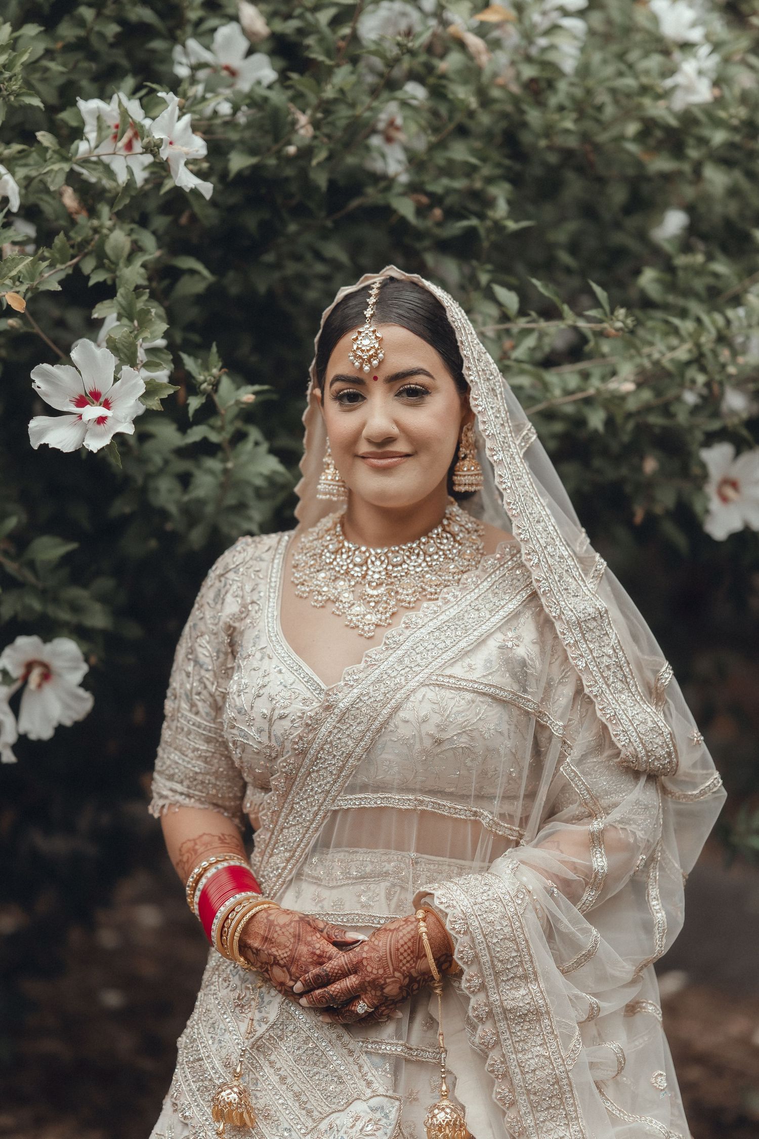 Bride in detailed ivory lehenga poses against flowering bushes in garden setting.