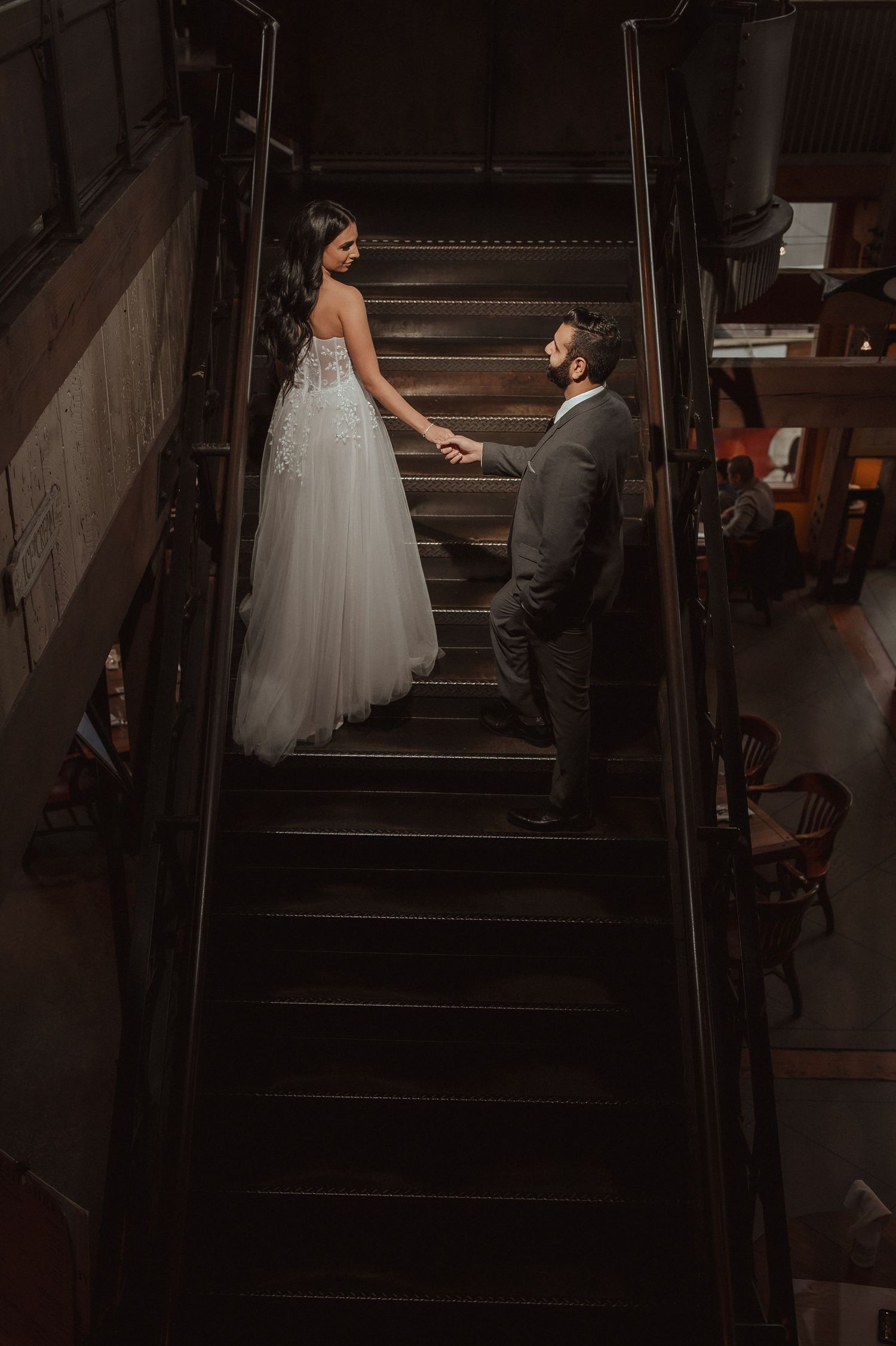 A person in a white wedding dress carefully descends dark stairs while being assisted from behind.