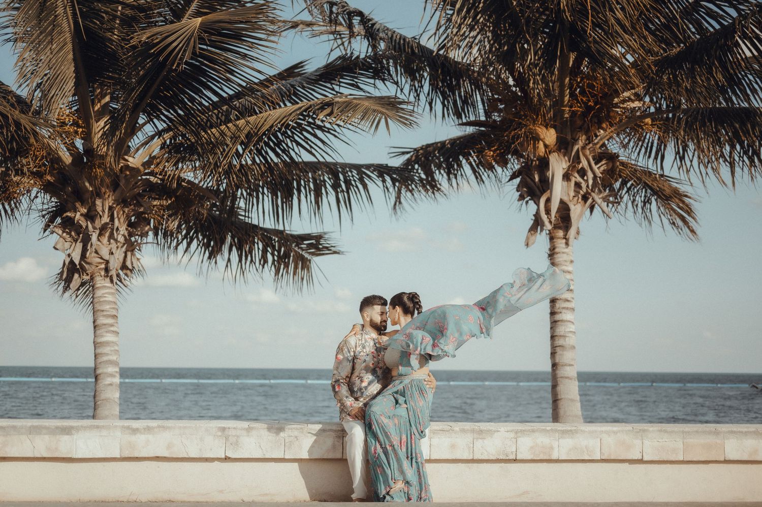 A couple shares romantic moments under swaying palm trees along a serene coastal beach at sunset.