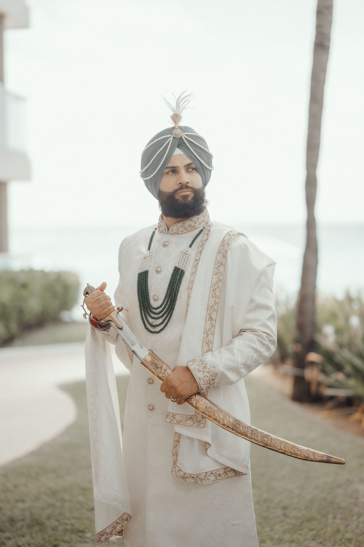 Groom in elegant white sherwani with gold embellishments and turban holds ceremonial sword at outdoor venue.