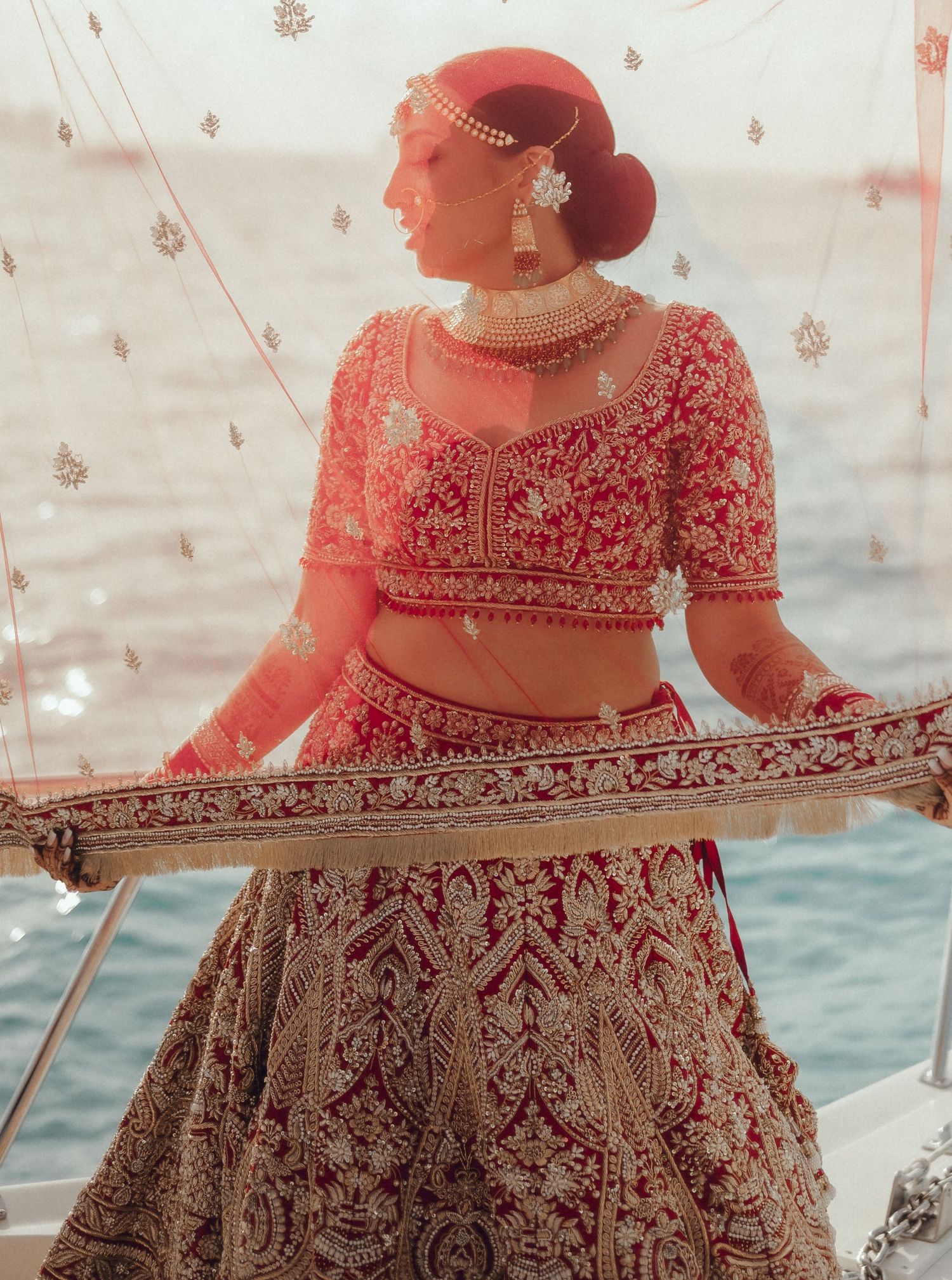 Person wearing an ornate red and gold embroidered lehenga photographed in sequence while posing by the water.