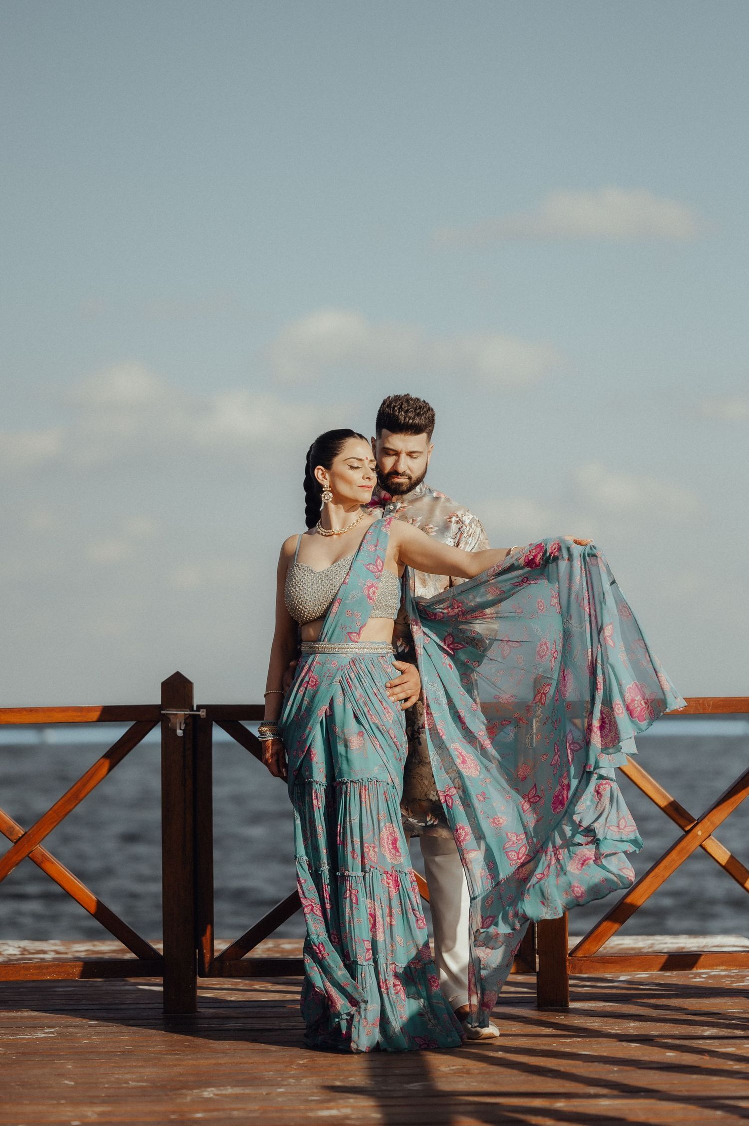 Couple in a light blue dress share romantic moments on a wooden pier by the ocean during sunset.