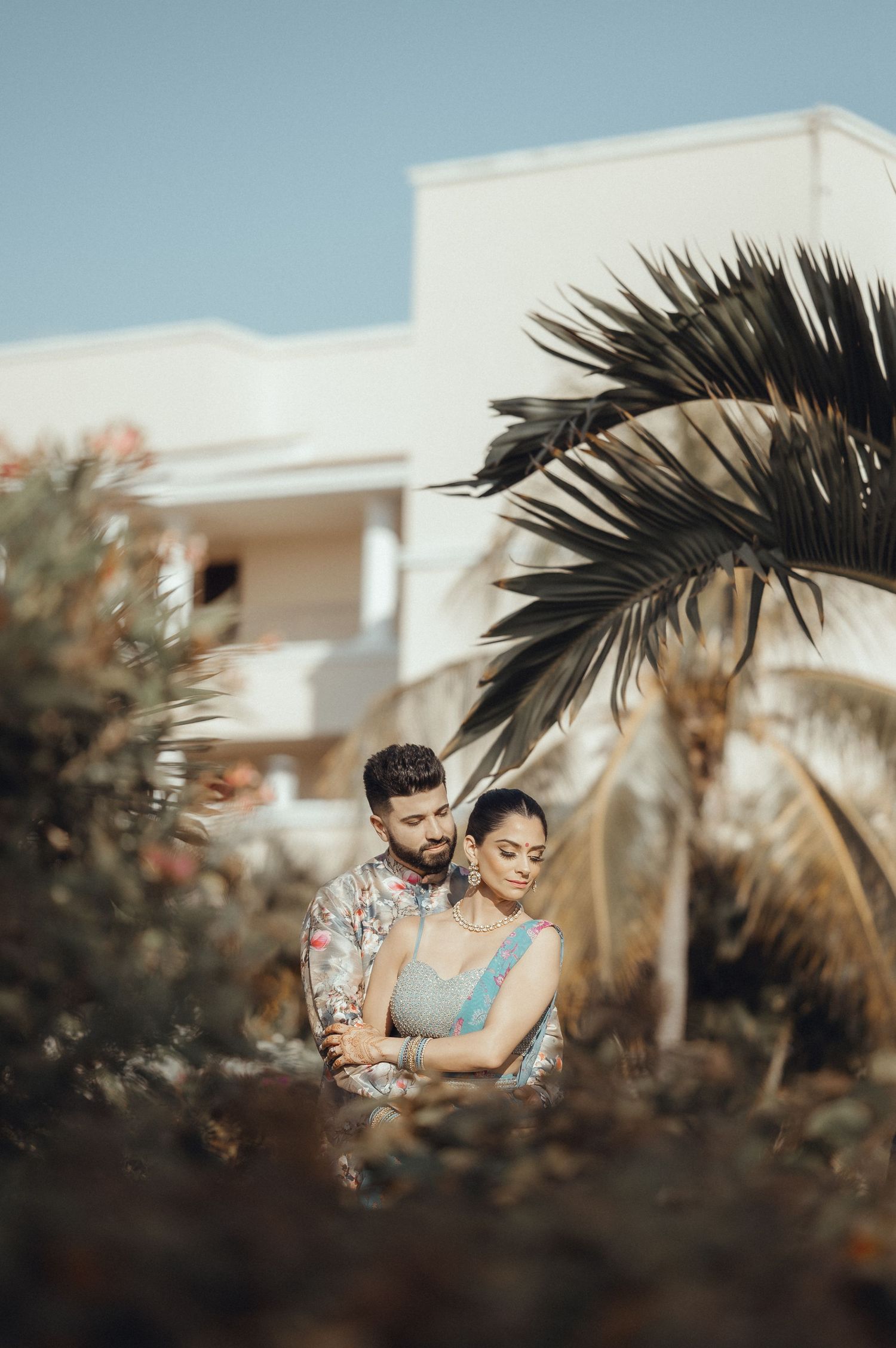 A romantic couple embraces in a warm golden light surrounded by tropical palm fronds and modern architecture.