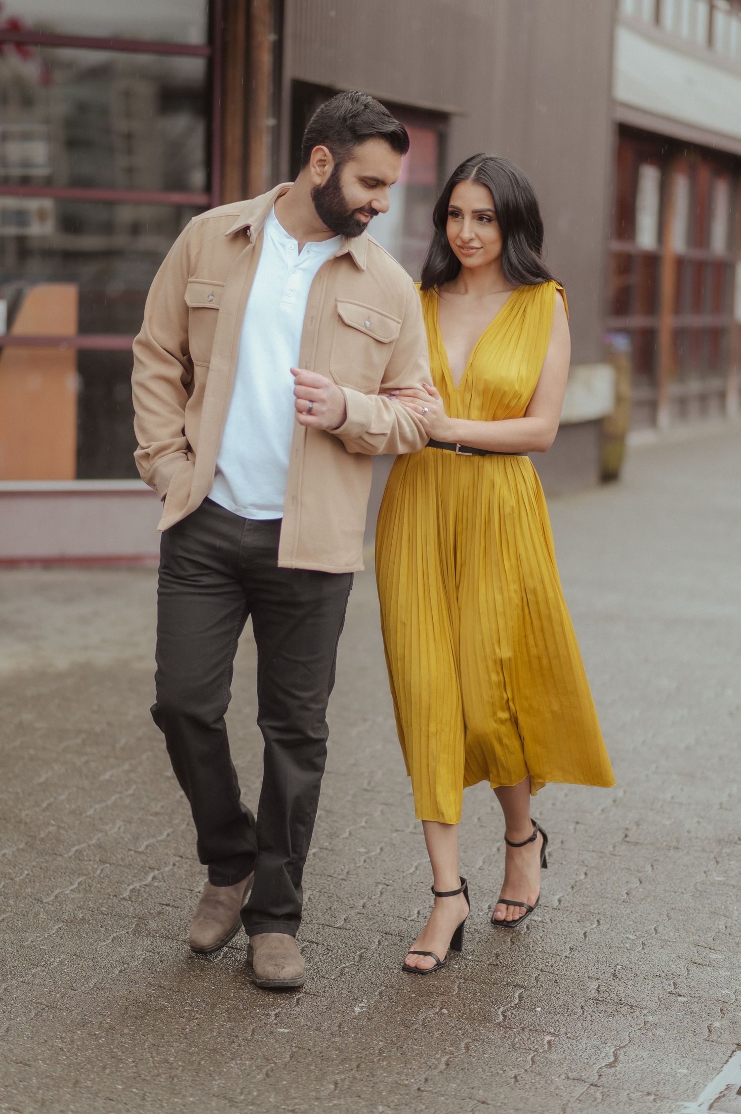 A couple walking together on stone pavement, with her wearing a vibrant yellow dress and him in a tan jacket.