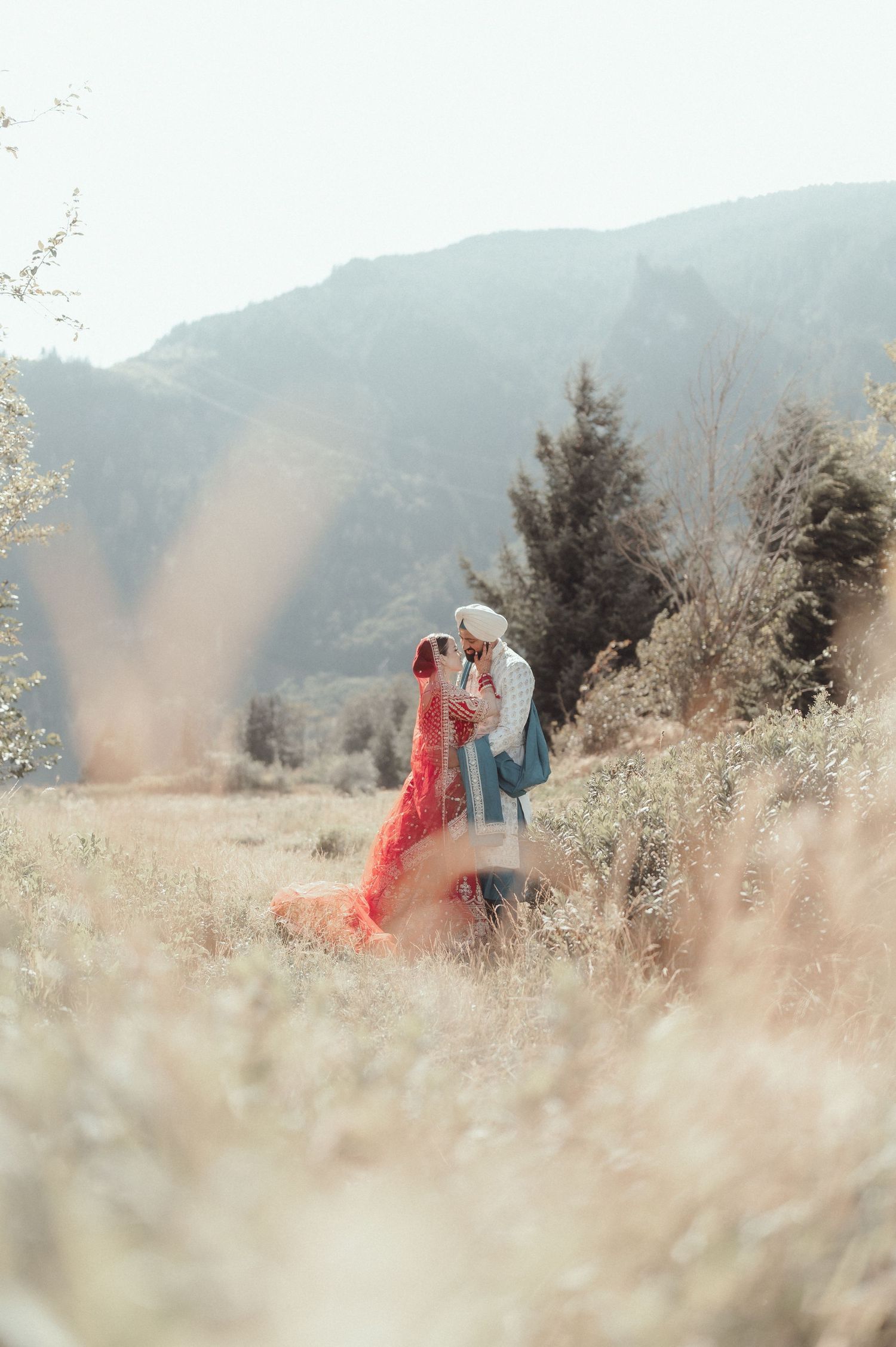 A romantic couple embraces in a dreamy meadow with soft sunlight and mountains in the background for their engagement photos.
