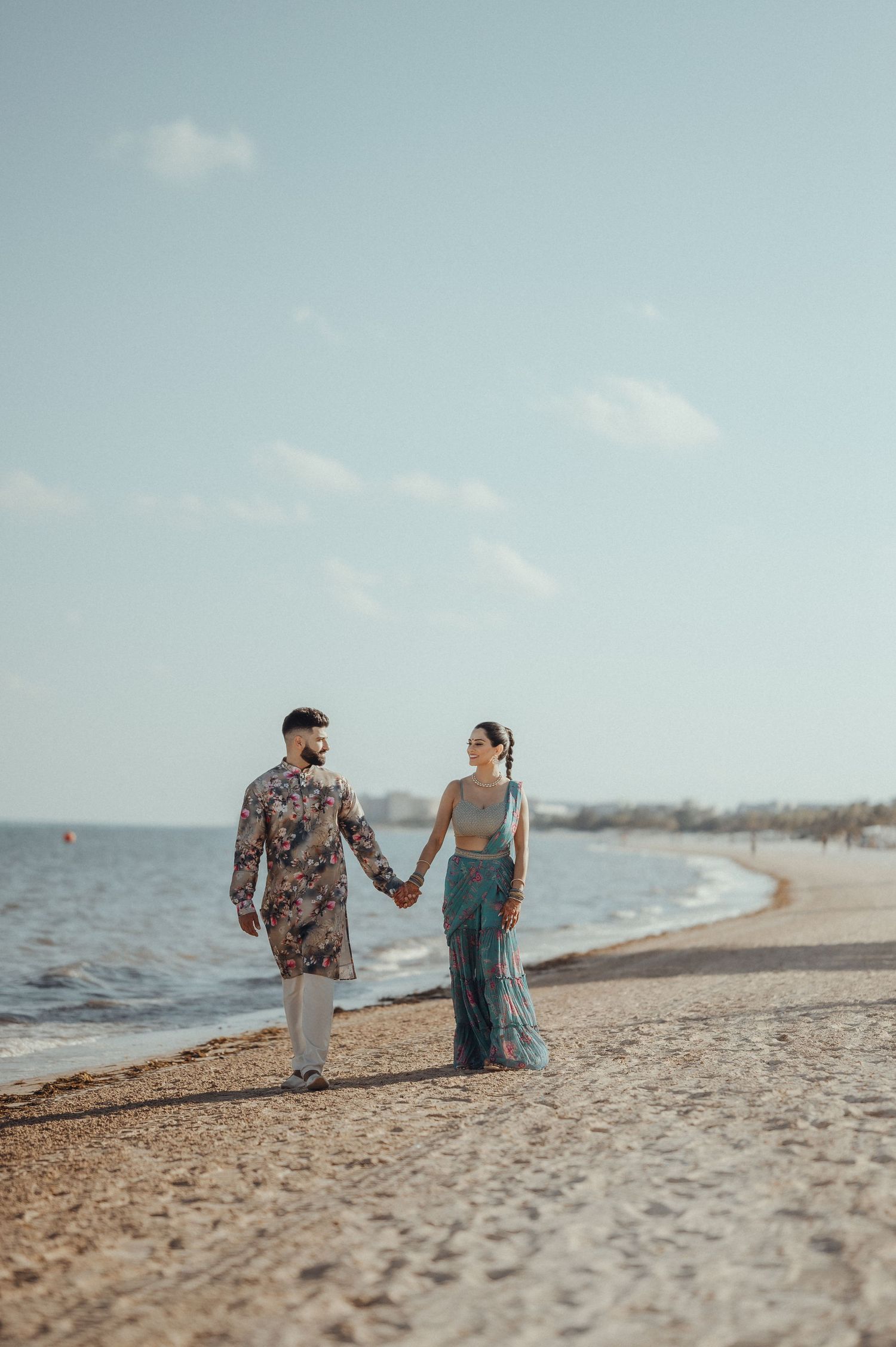 A couple walking hand in hand along a sandy beach during sunset with calm waters and soft blue sky in the background.