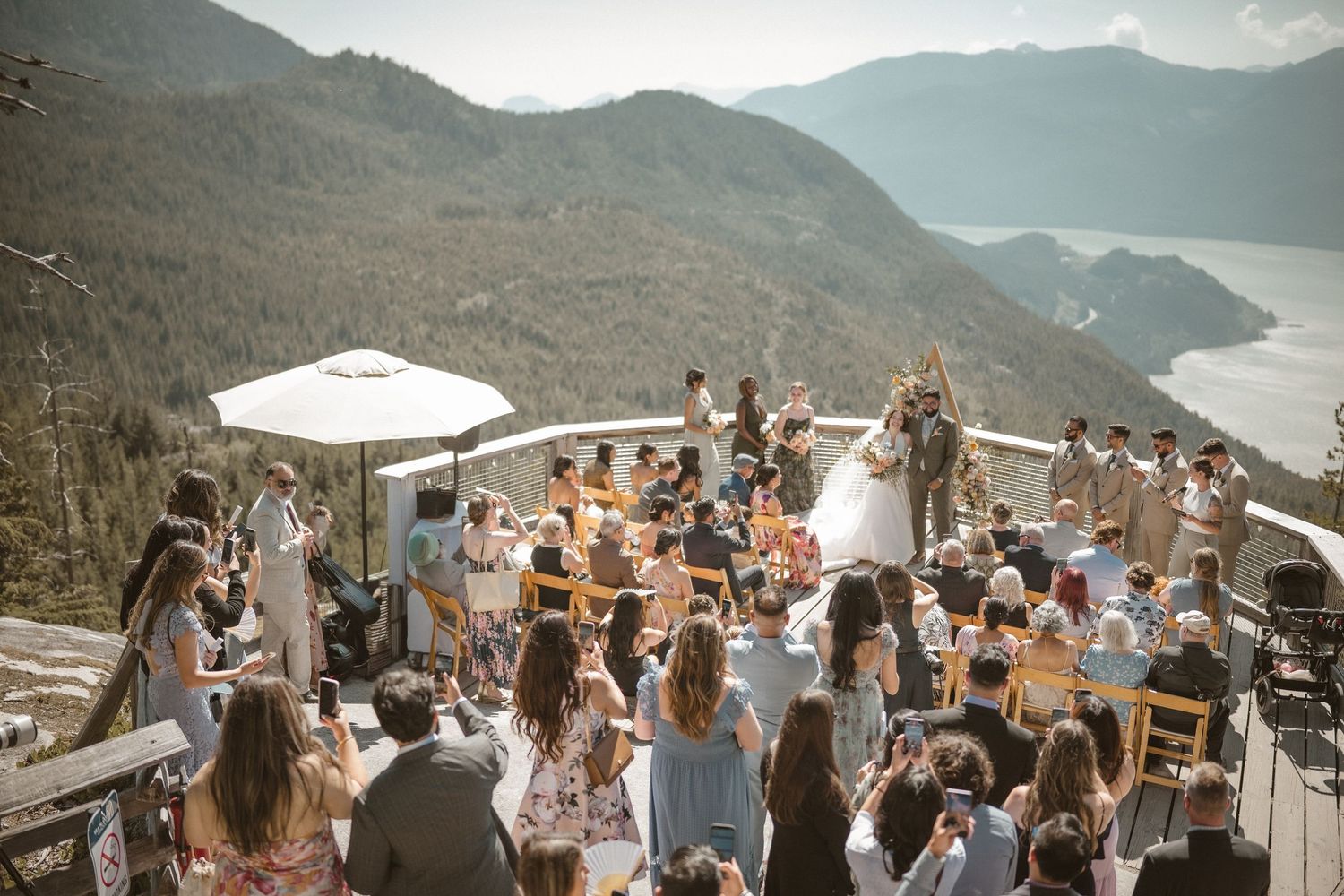 Wedding guests celebrate outdoors with mountains and lake views in the background on a sunny day.
