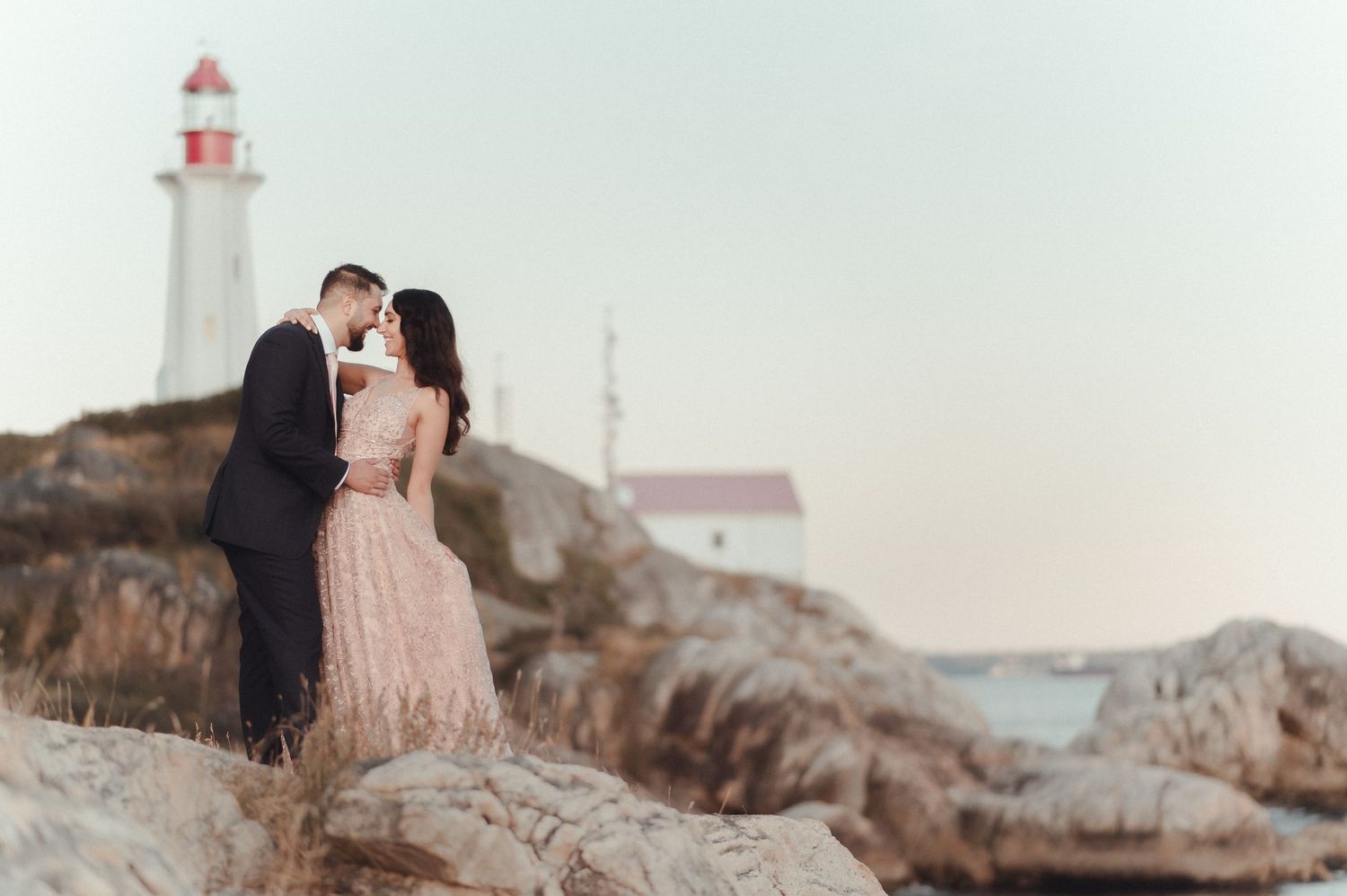 Couple embracing on coastal cliff with lighthouse in background during sunset.