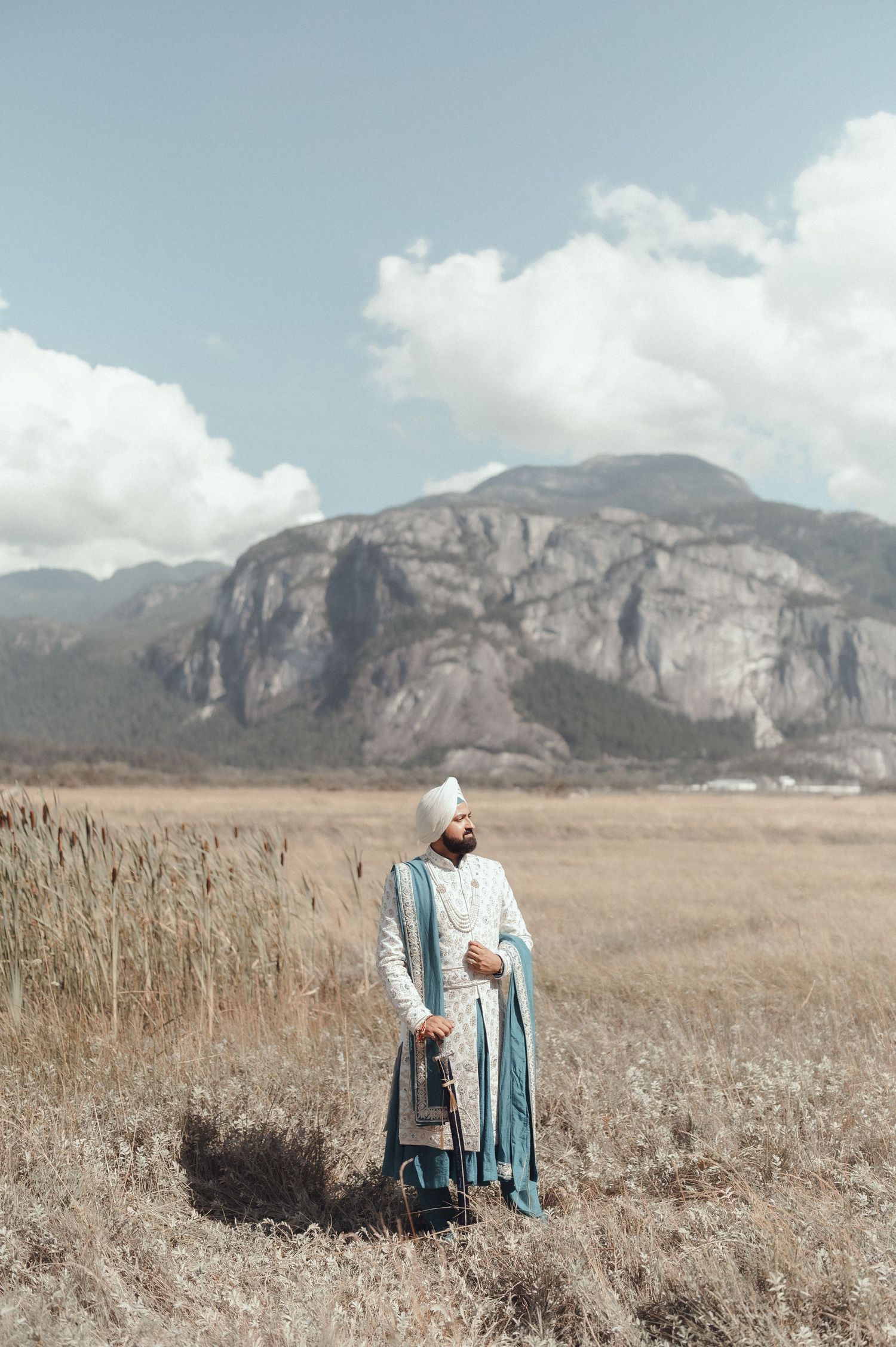 A couple stands together in a golden field with rugged mountain cliffs and cloudy skies in the backdrop.