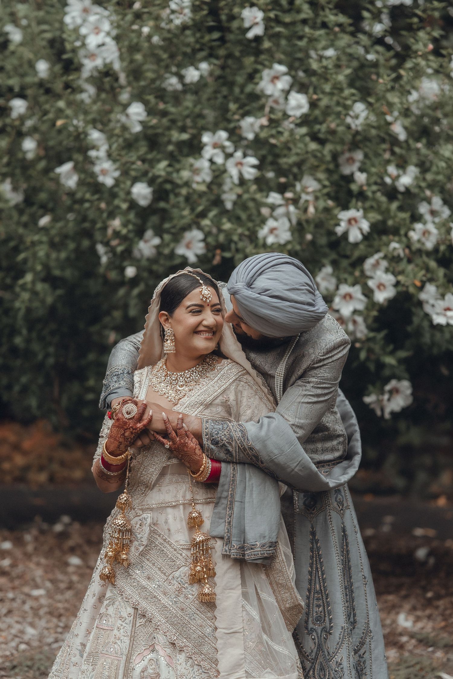 Joyful wedding couple in traditional Indian attire posing among white flowering trees.