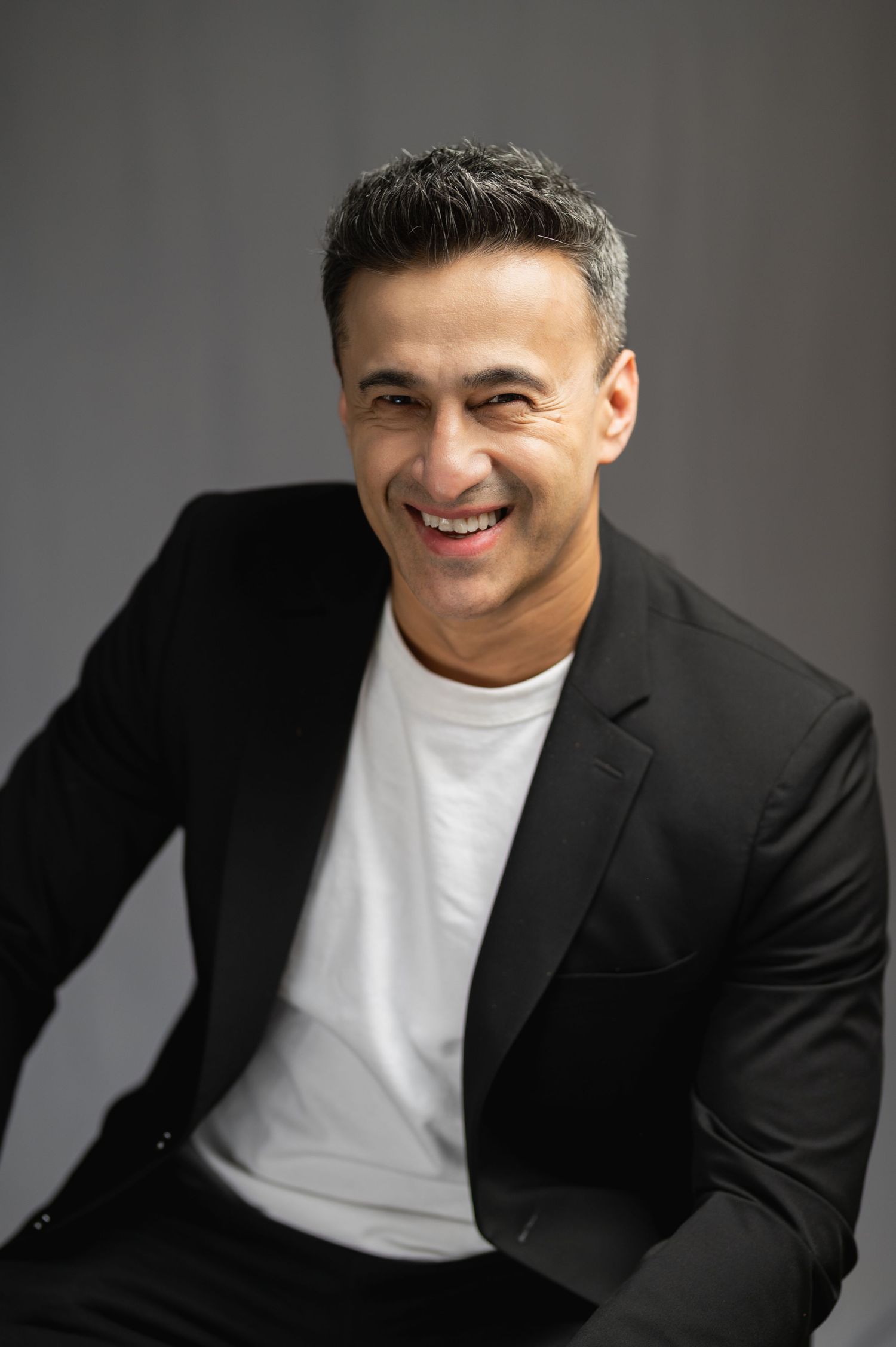 Smiling middle-aged man in black blazer and white t-shirt posing confidently against a gray background.