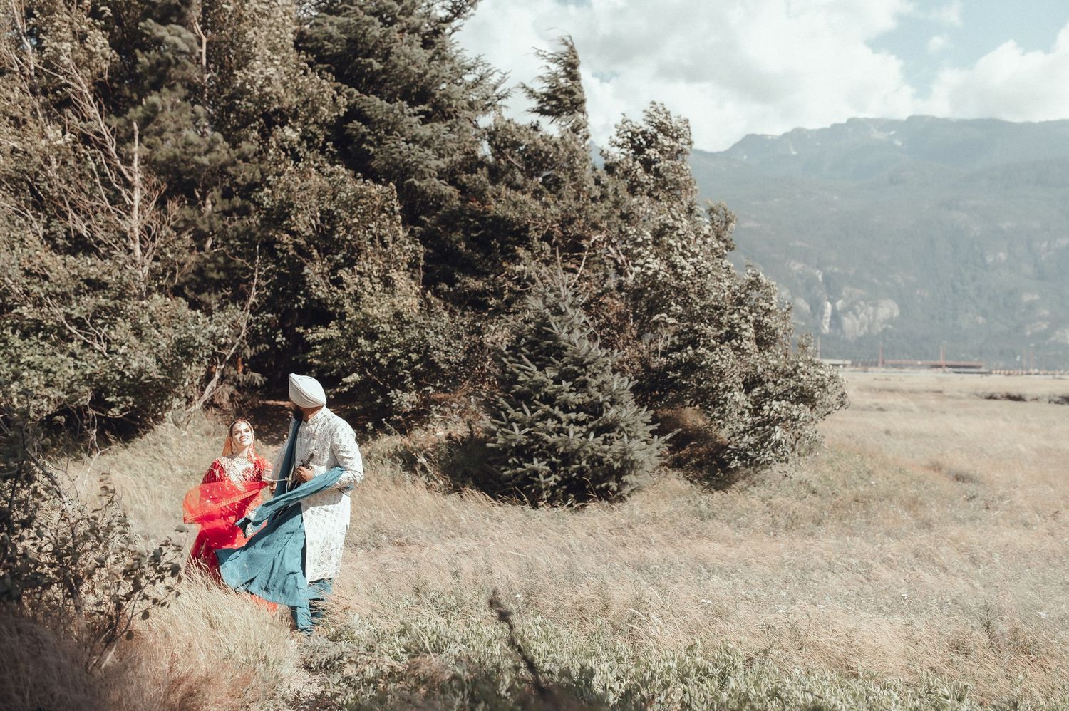 Two people walk together through a scenic mountain meadow with evergreen trees in the distance on a soft overcast day.