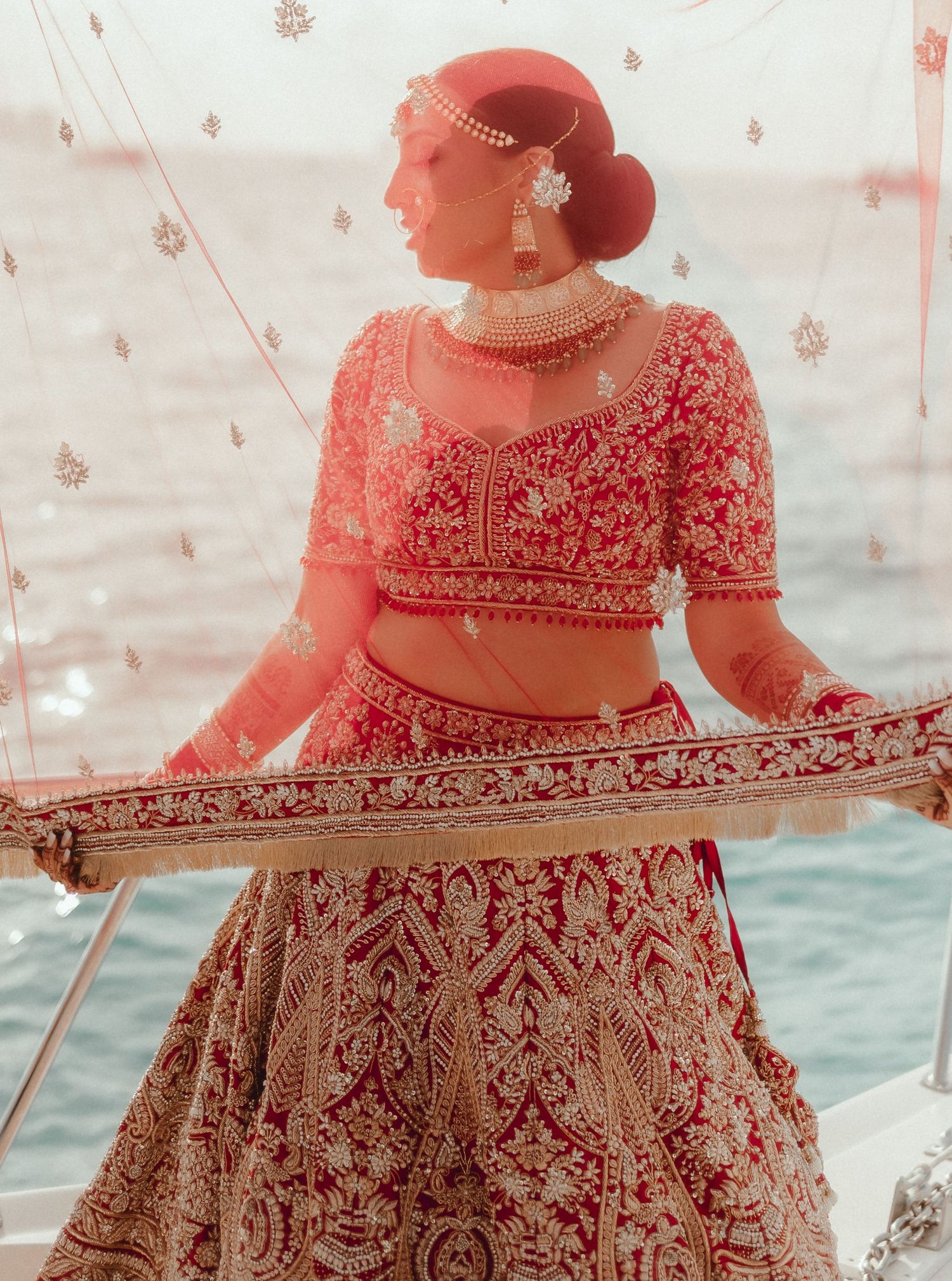 A bride in traditional red and gold Indian lehenga posing with ornate details by the water.