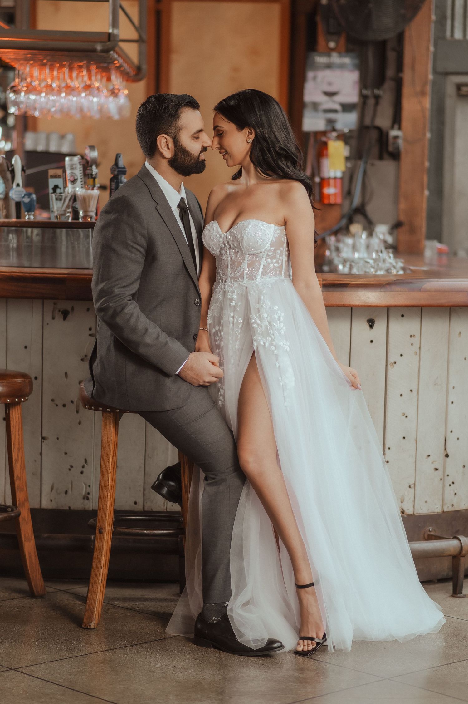 A romantic couple shares an intimate moment at a rustic bar, with the bride wearing a flowing white gown with high slit.