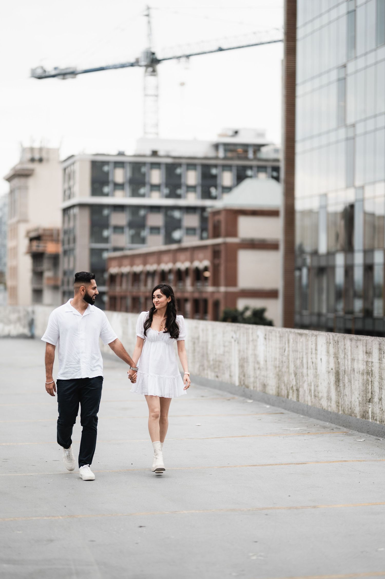 Couple in white outfits walking along concrete wall with urban backdrop.