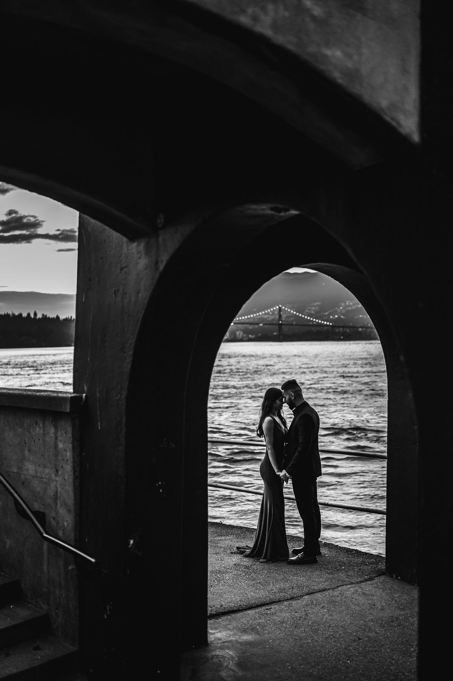 Silhouetted couple embracing under concrete arches with dramatic sunset and bridge view.