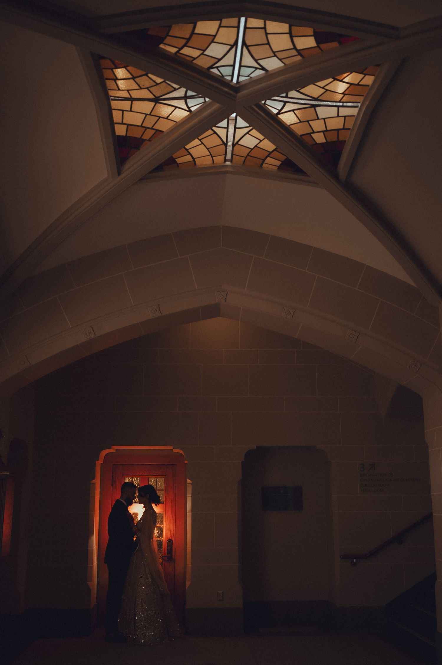 A dark spooky hallway with arched ceilings and stained glass windows transitions into a dimly lit orange doorway.