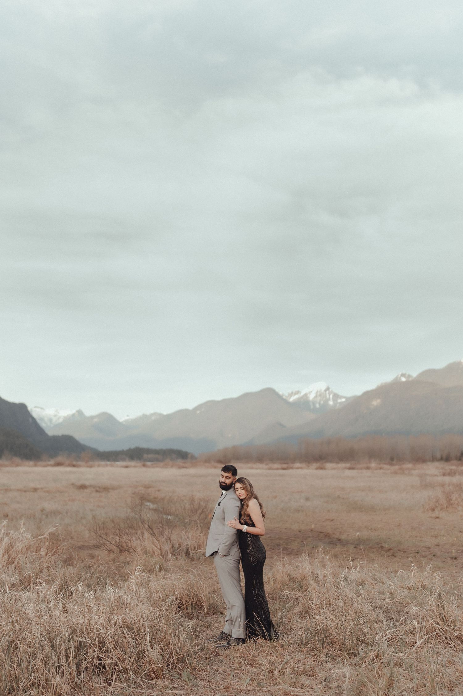 Couple in formal wear sharing intimate moment in brown grass field with mountains behind.