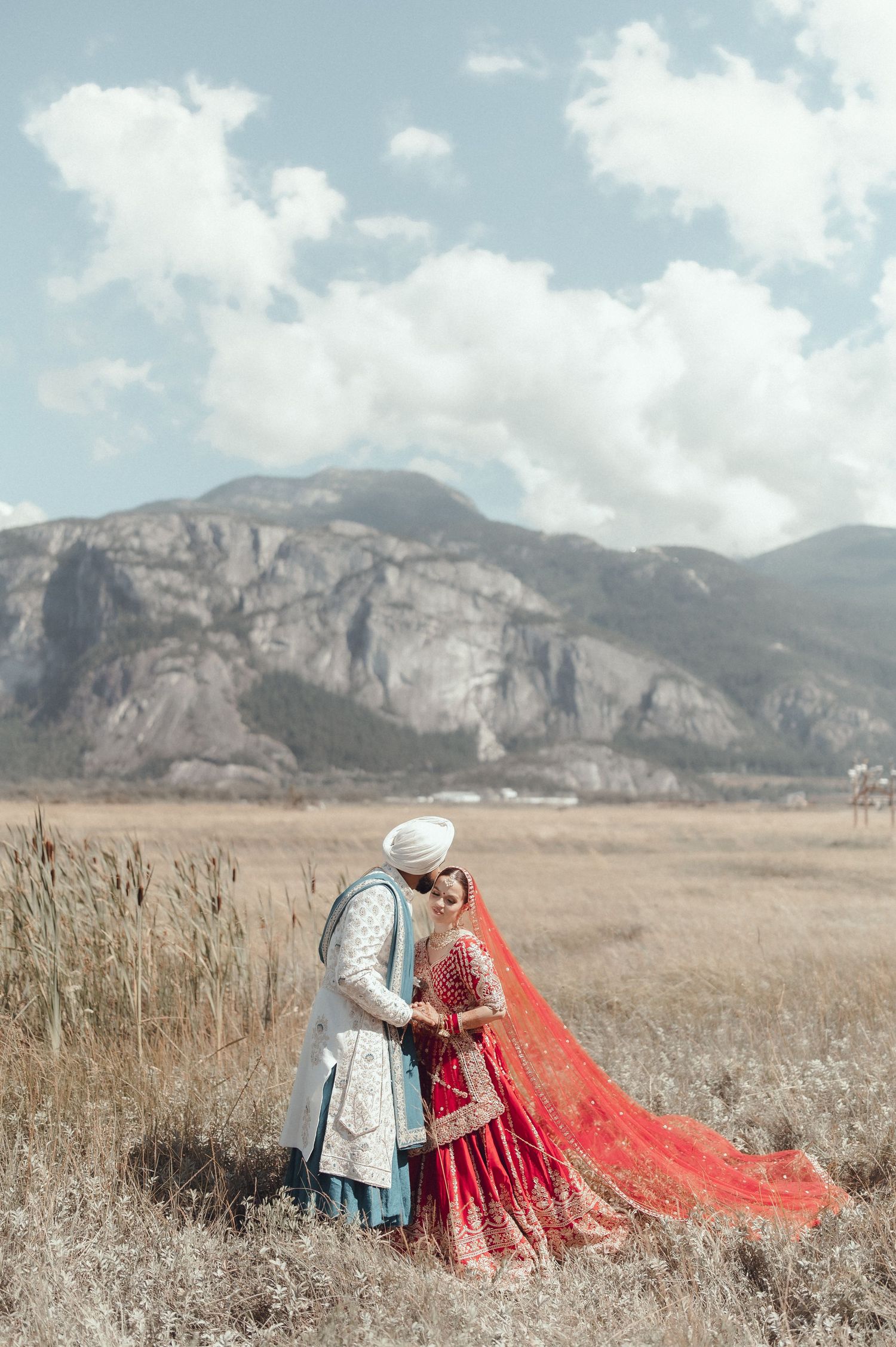 A couple in traditional wedding attire poses in a field with dramatic mountain cliffs and clouds in the background.