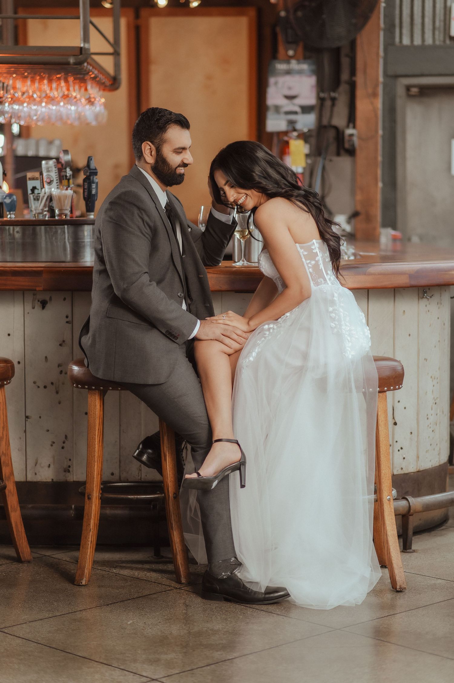 A couple in wedding attire shares an intimate moment while sitting at a rustic bar counter.
