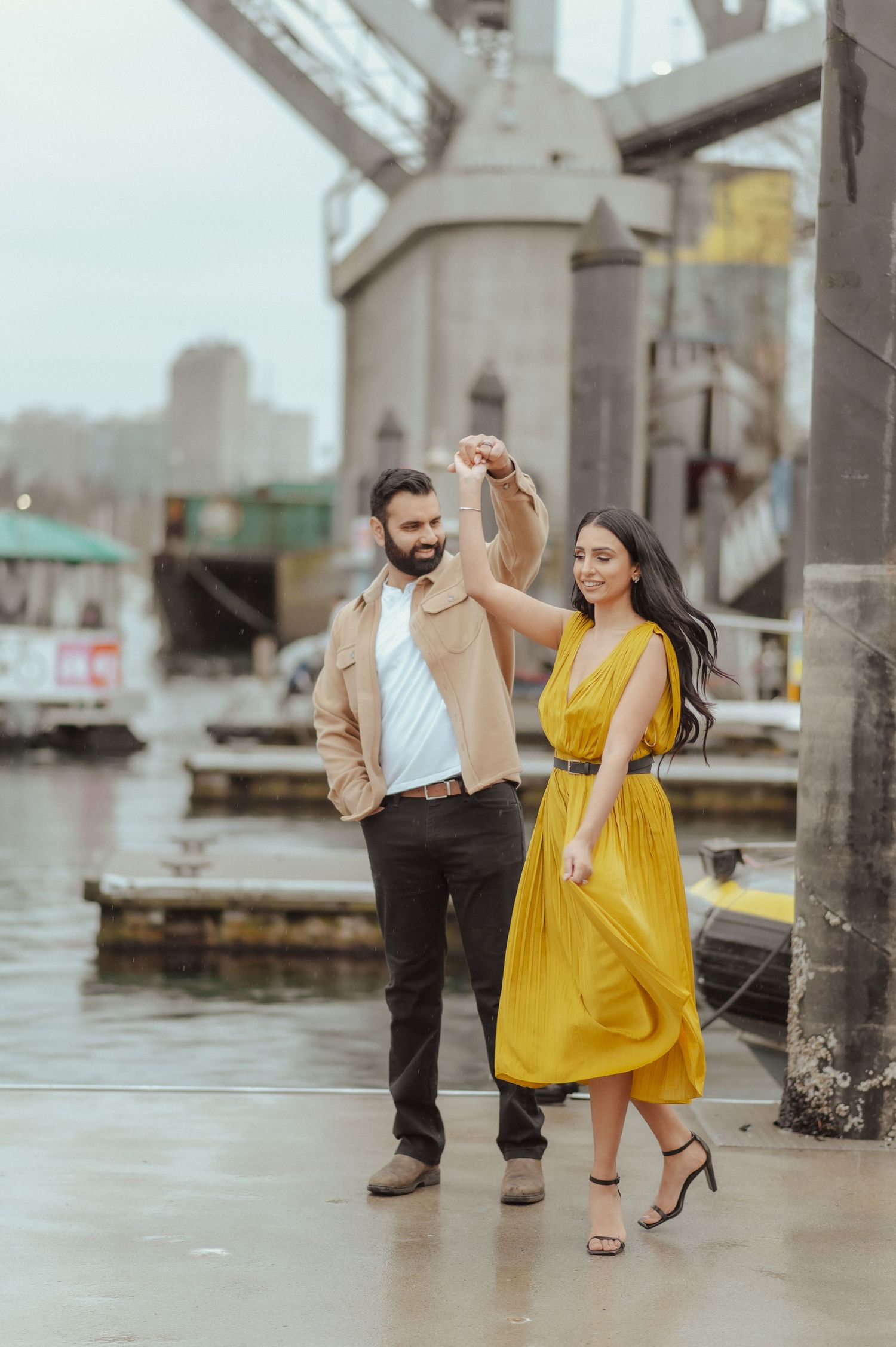 A couple dances playfully on a wet dock with industrial architecture in the background.