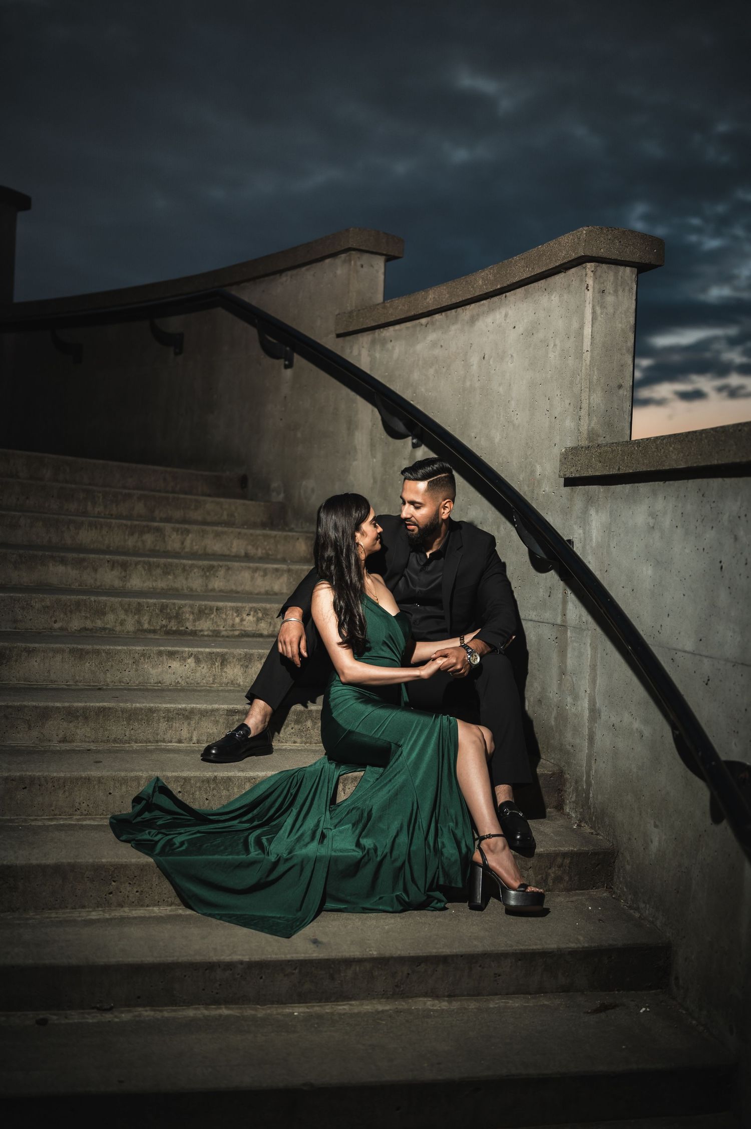 Couple in formal attire posing dramatically on concrete steps at dusk.
