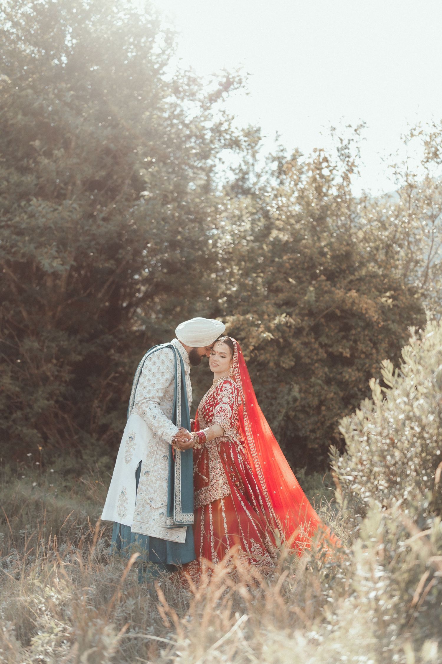 A couple in traditional wedding attire shares an intimate moment in a sunlit meadow with soft golden bokeh.