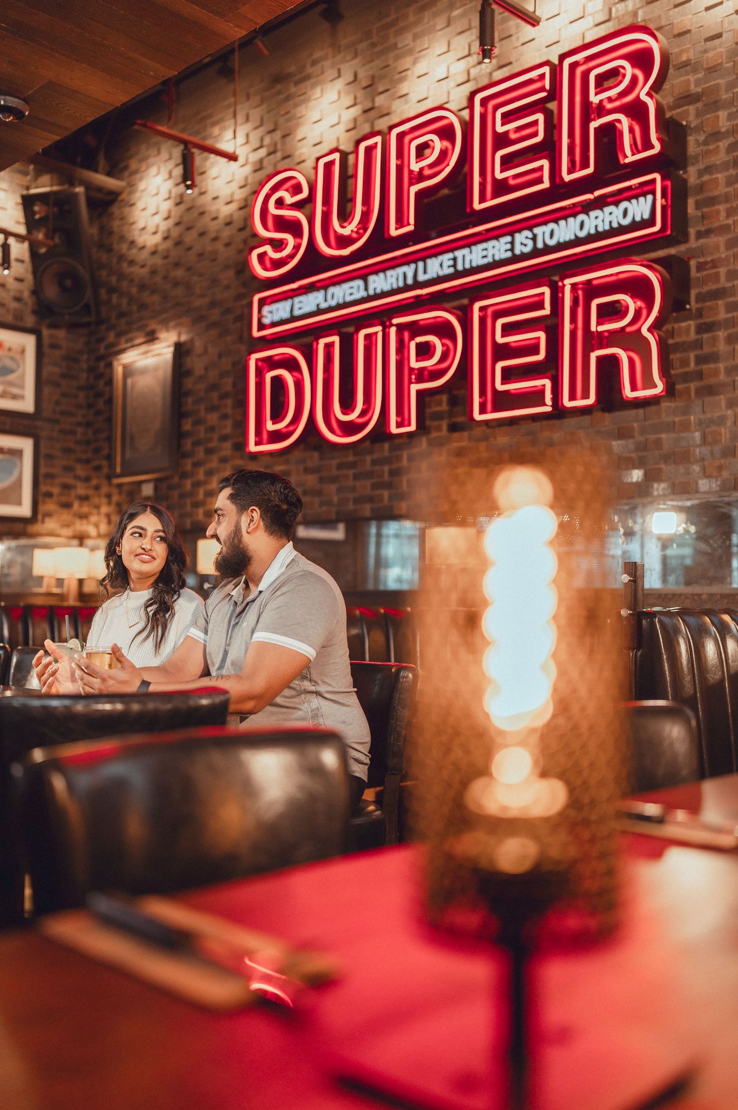Couple sharing drinks at a bar with neon 'Super Duper' sign glowing on brick wall.