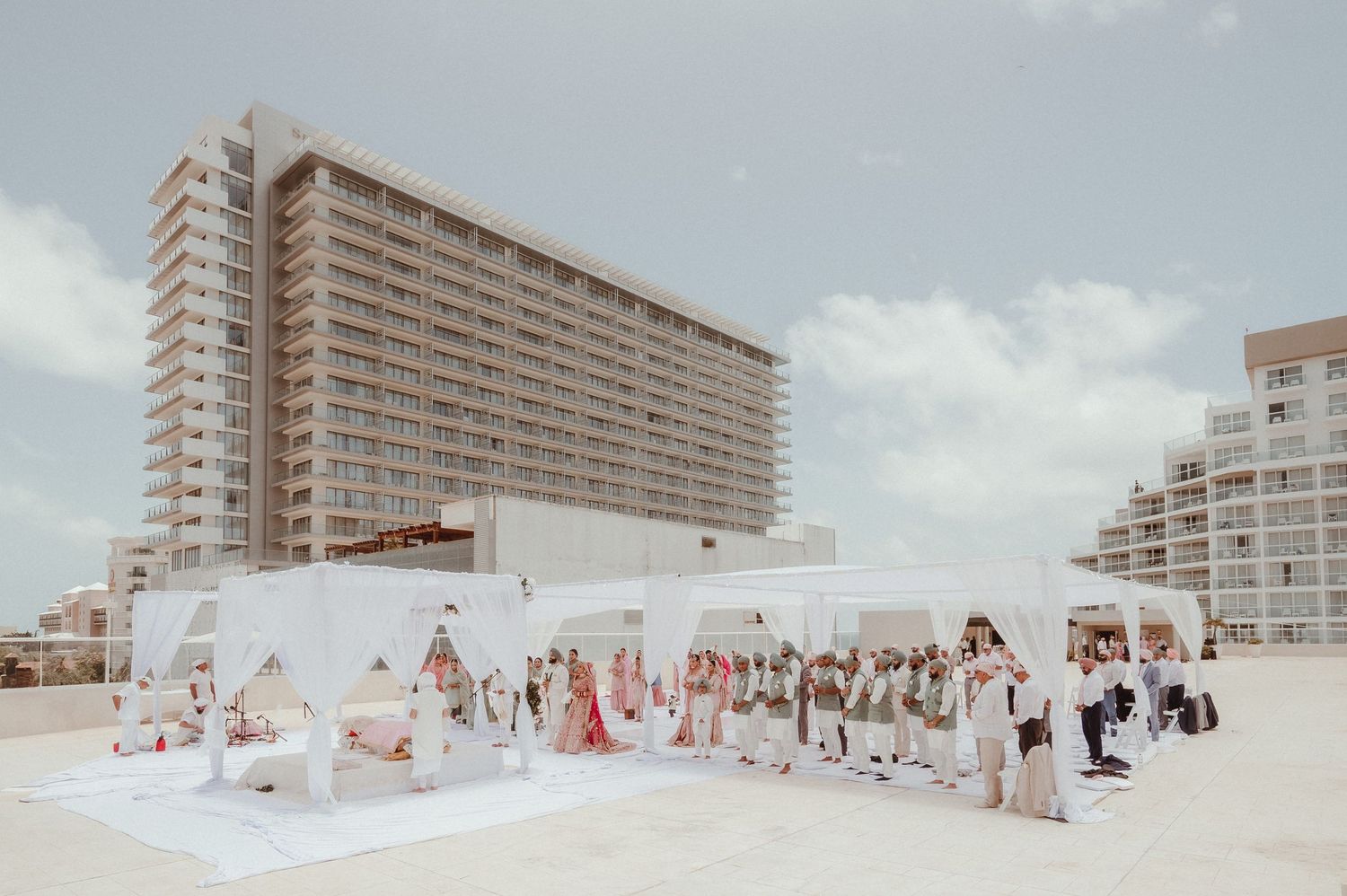 Panoramic view of a coastal beach wedding setup with white tents and high-rise buildings in background.