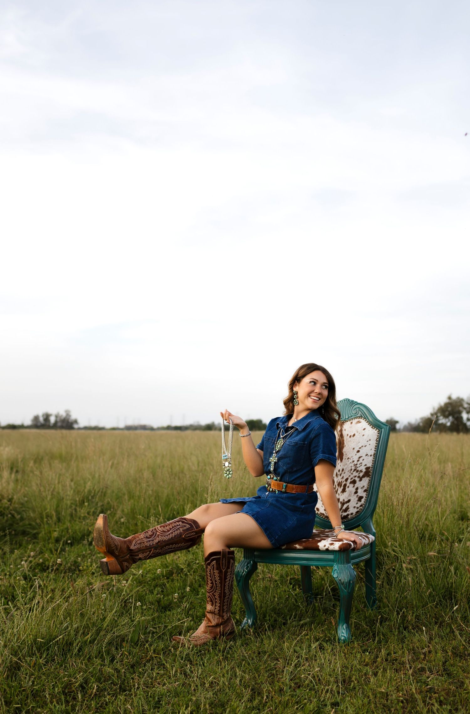 Woman in blue dress and cowboy boots sits on teal chair in open field, smiling and holding beaded necklace.