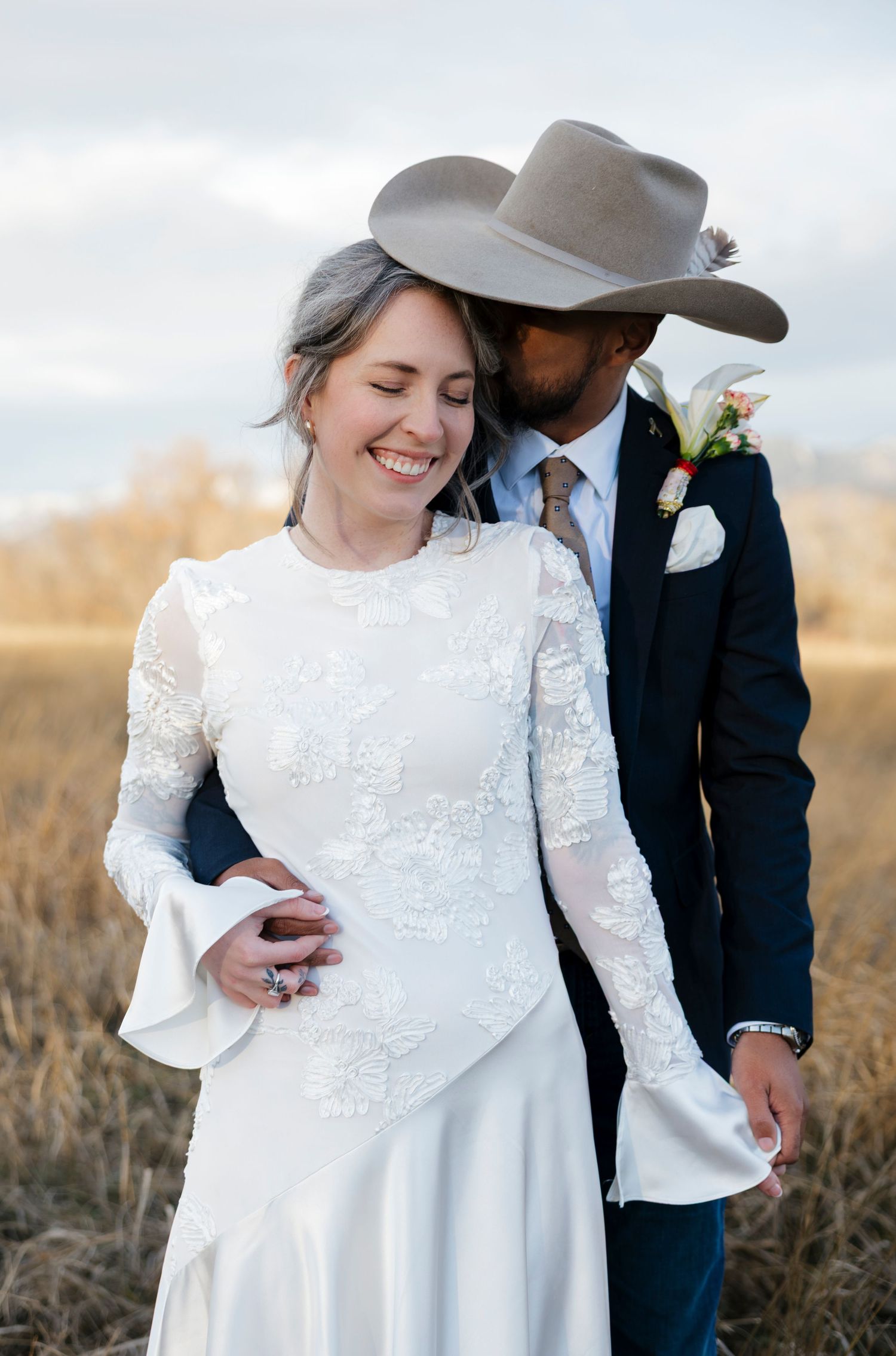 A romantic wedding couple embraces in a field during golden hour, with the bride wearing a white lace dress.