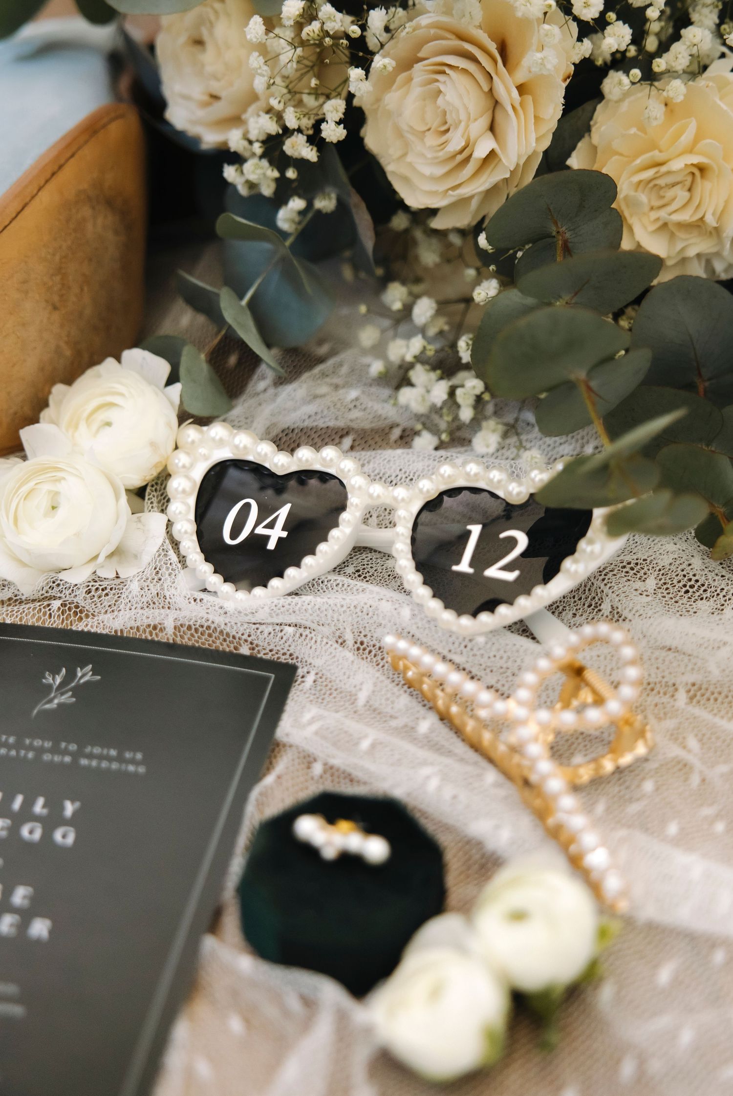 Wedding details featuring black stationery, pearls, and cream roses with eucalyptus leaves.
