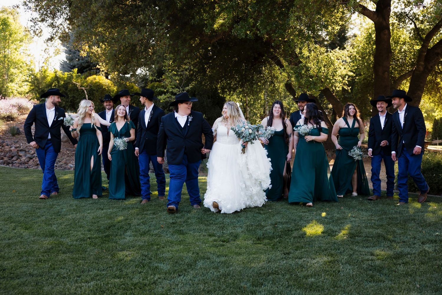 Wedding party in navy and teal attire poses together on green lawn during outdoor celebration.