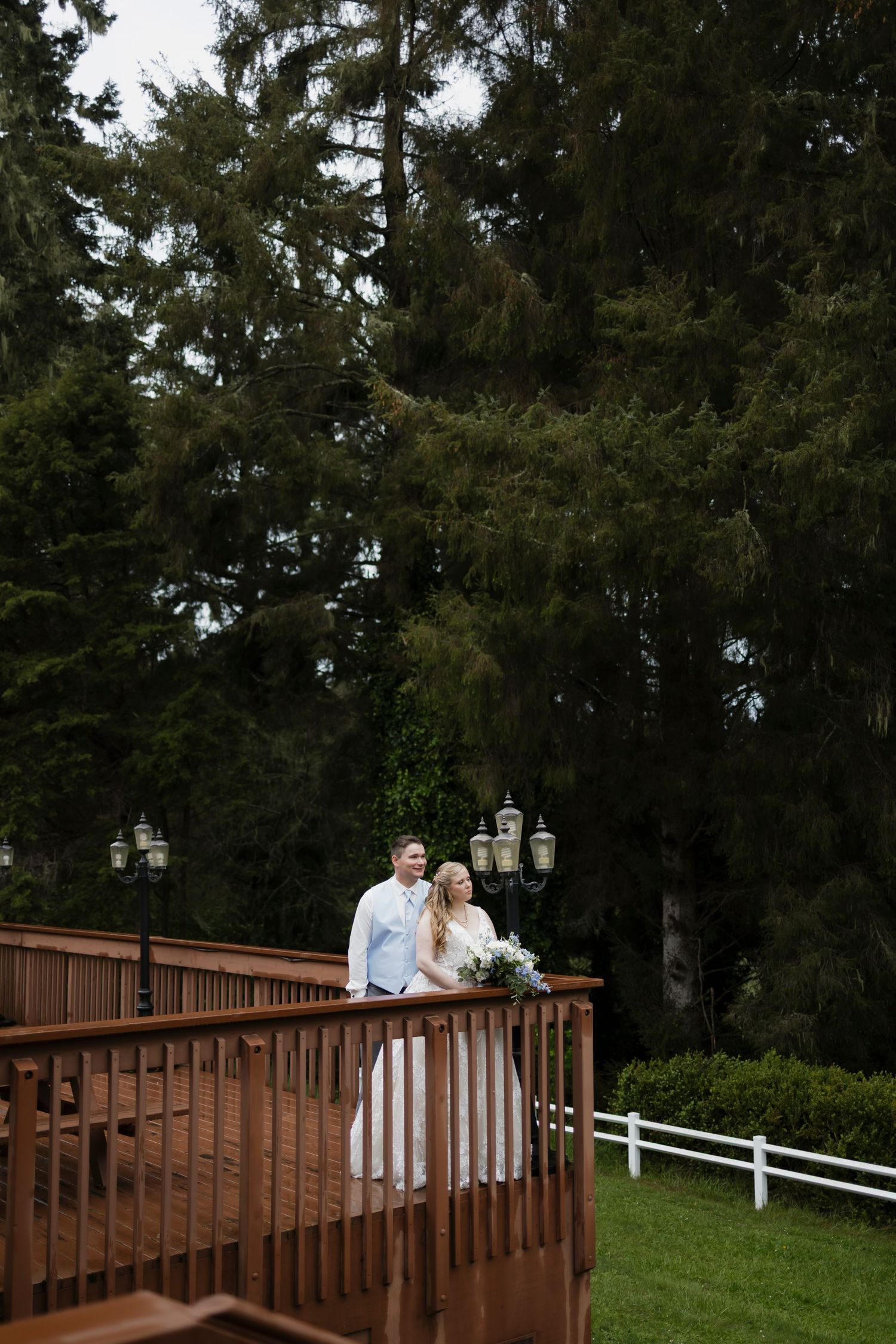 A bride and groom pose together on a wooden deck surrounded by tall evergreen trees at an outdoor wedding venue.