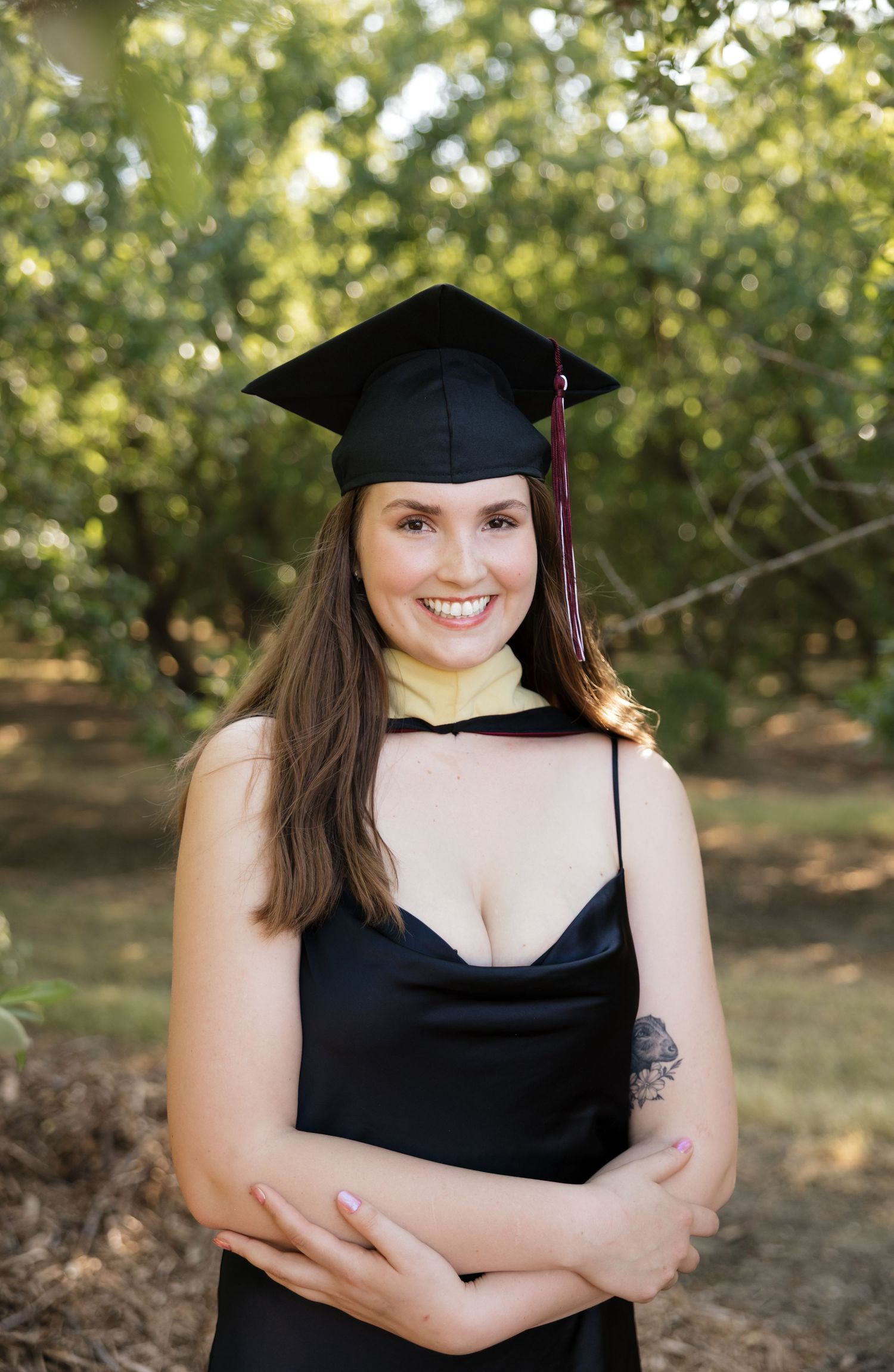 Graduate wearing black cap and gown poses against leafy background.
