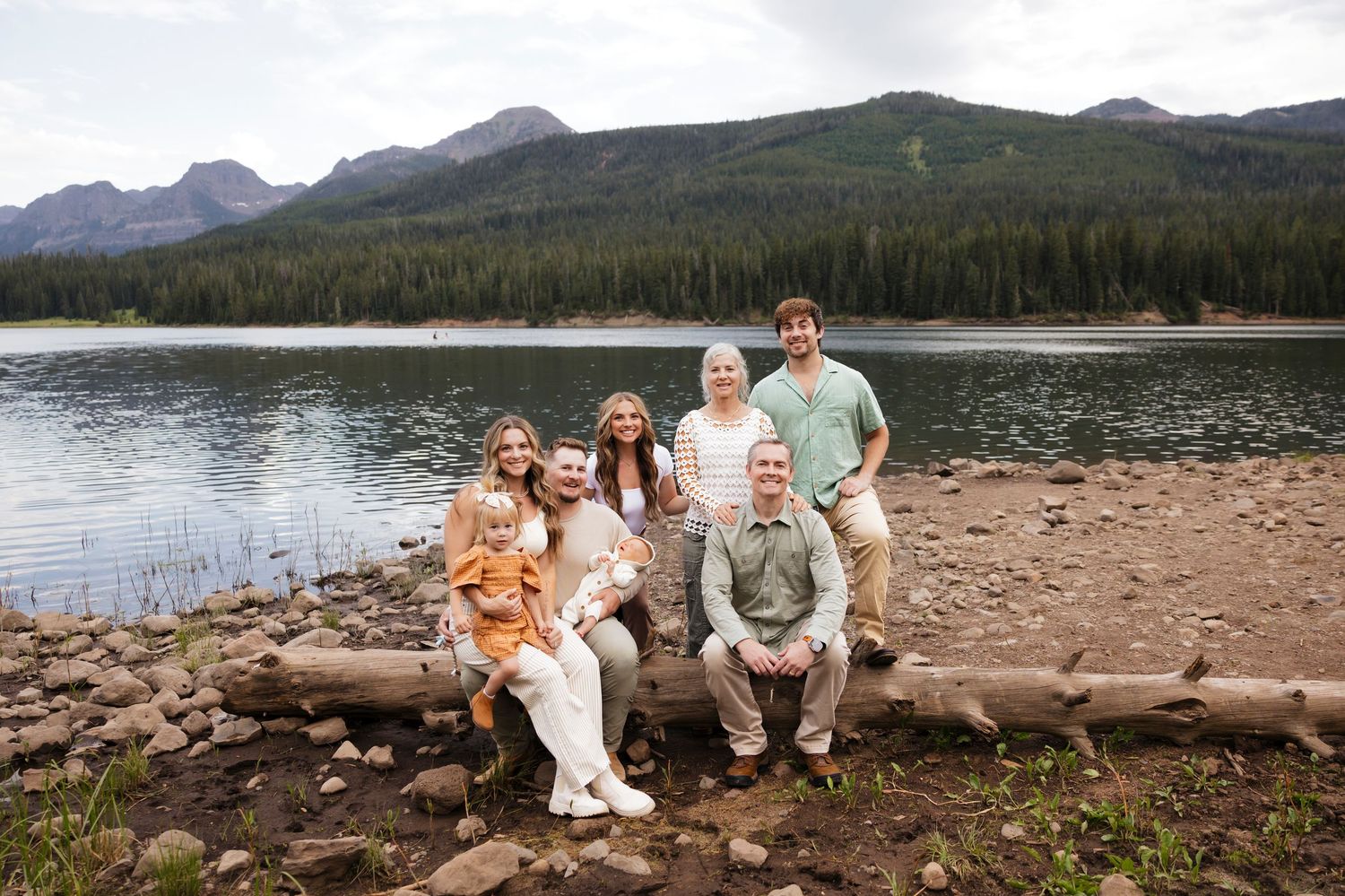 Family of six posing by a mountain lake surrounded by pine trees and rocky peaks in a scenic outdoor setting.
