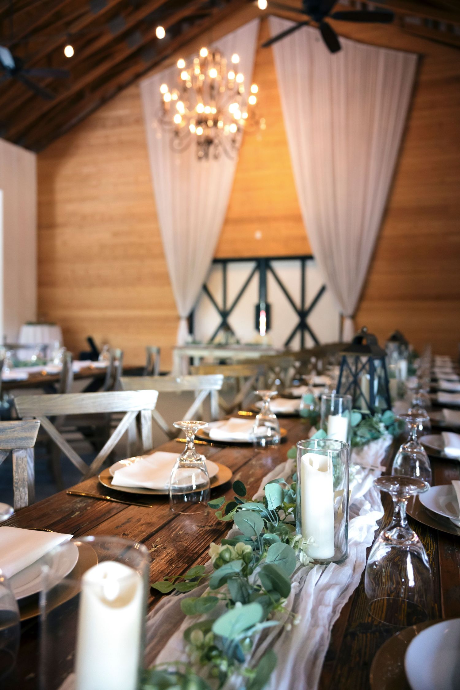 Rustic wedding reception table setup with wooden tables, chandeliers and white draping.