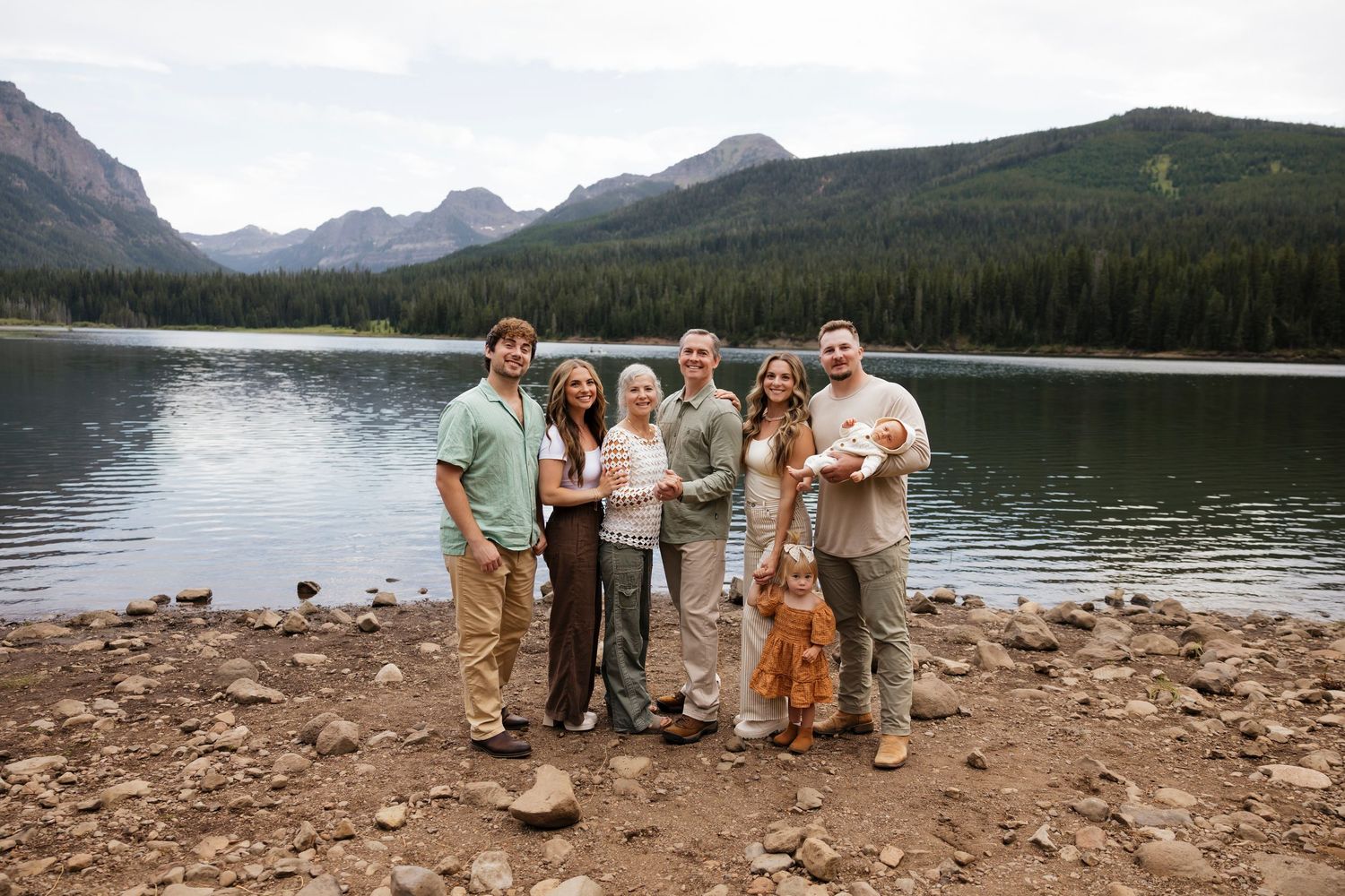 Group of seven people smiling by a mountain lake with forested peaks in the background on a cloudy day.