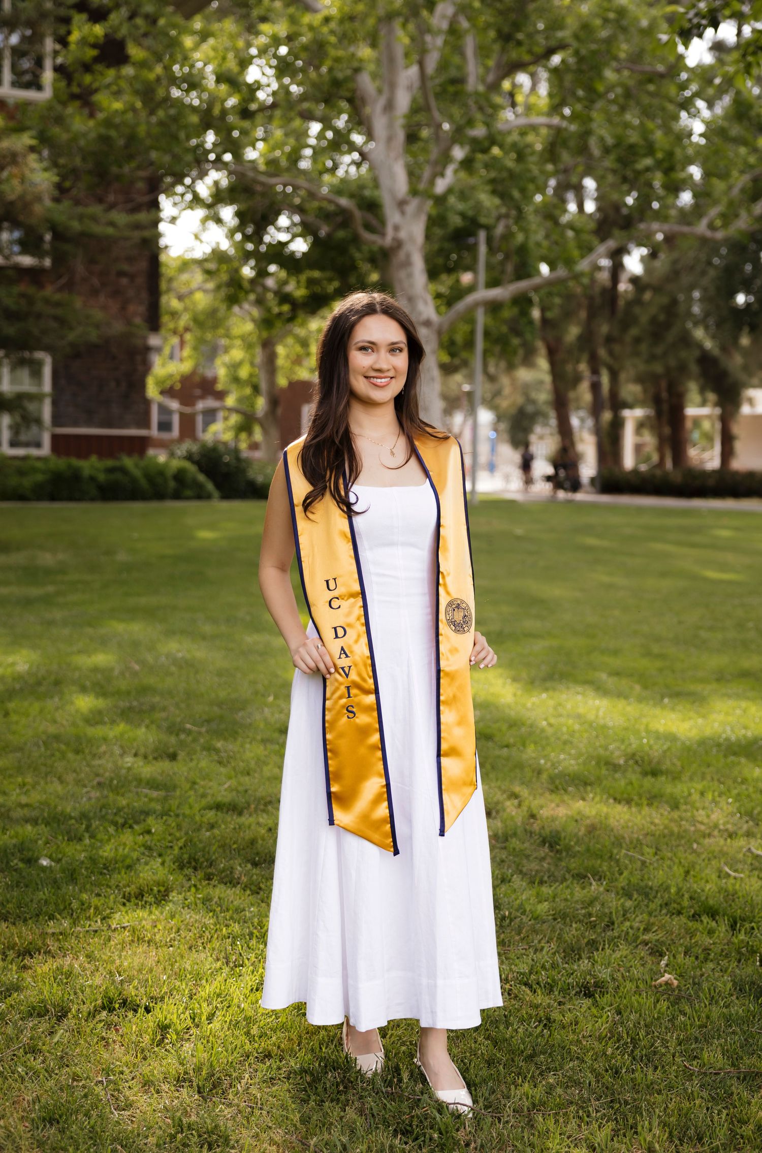 Graduate wearing a white dress and gold sash stands on grass in front of brick building.