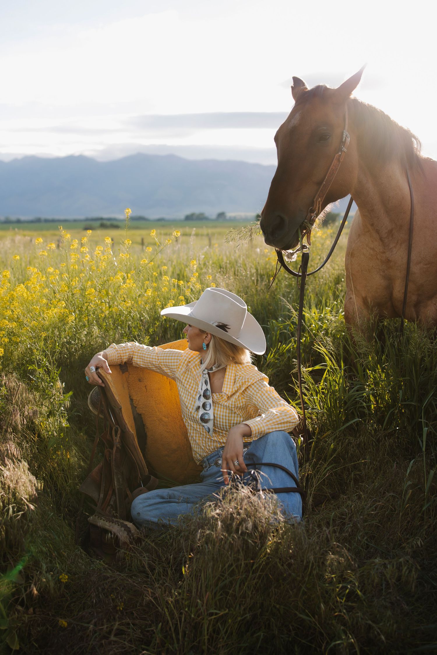 A cowgirl in a white hat and yellow plaid shirt sits in a wildflower field beside a chestnut horse at golden hour.
