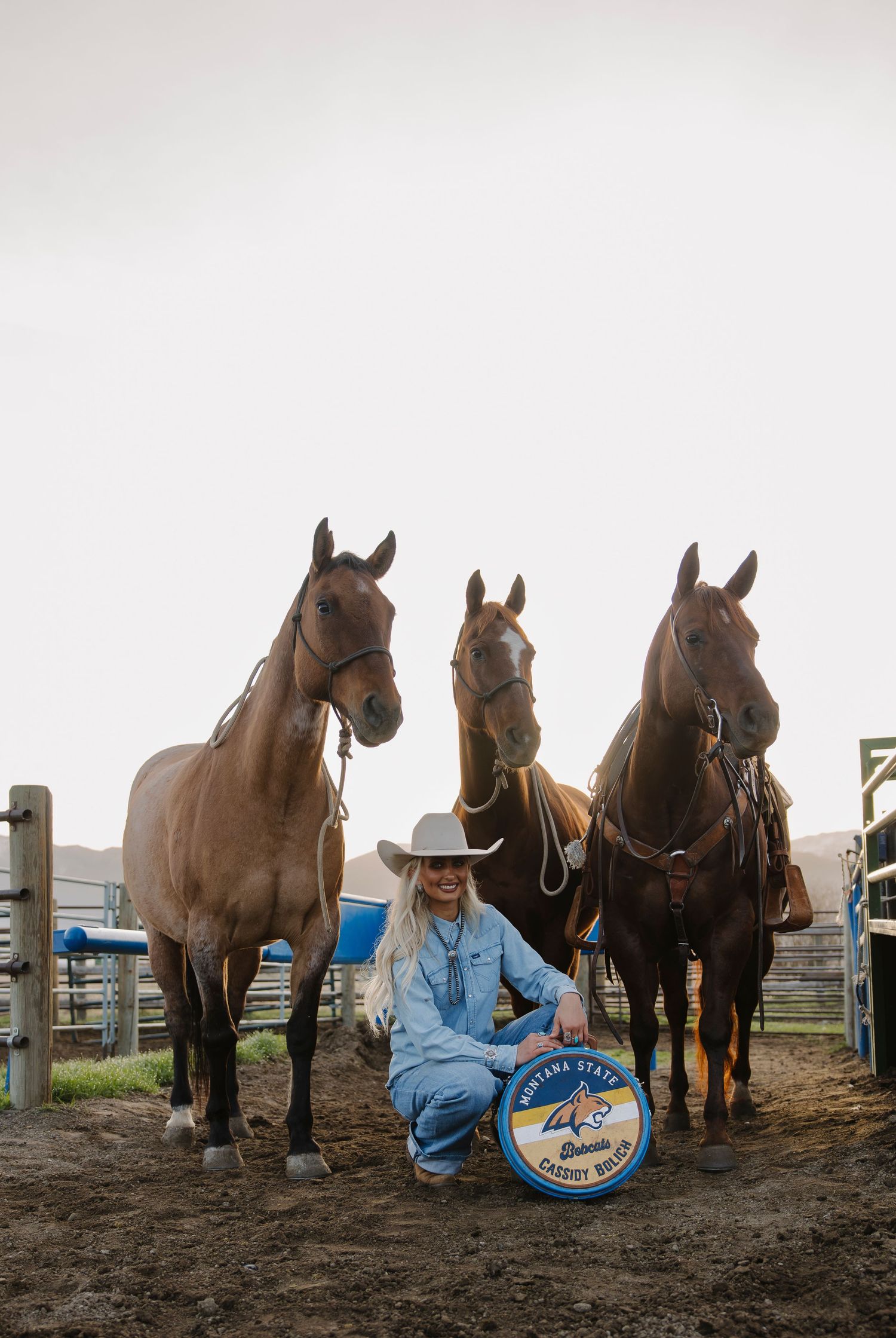 Woman in cowboy hat kneels with Montana State award plaque in front of three brown horses at a ranch corral.