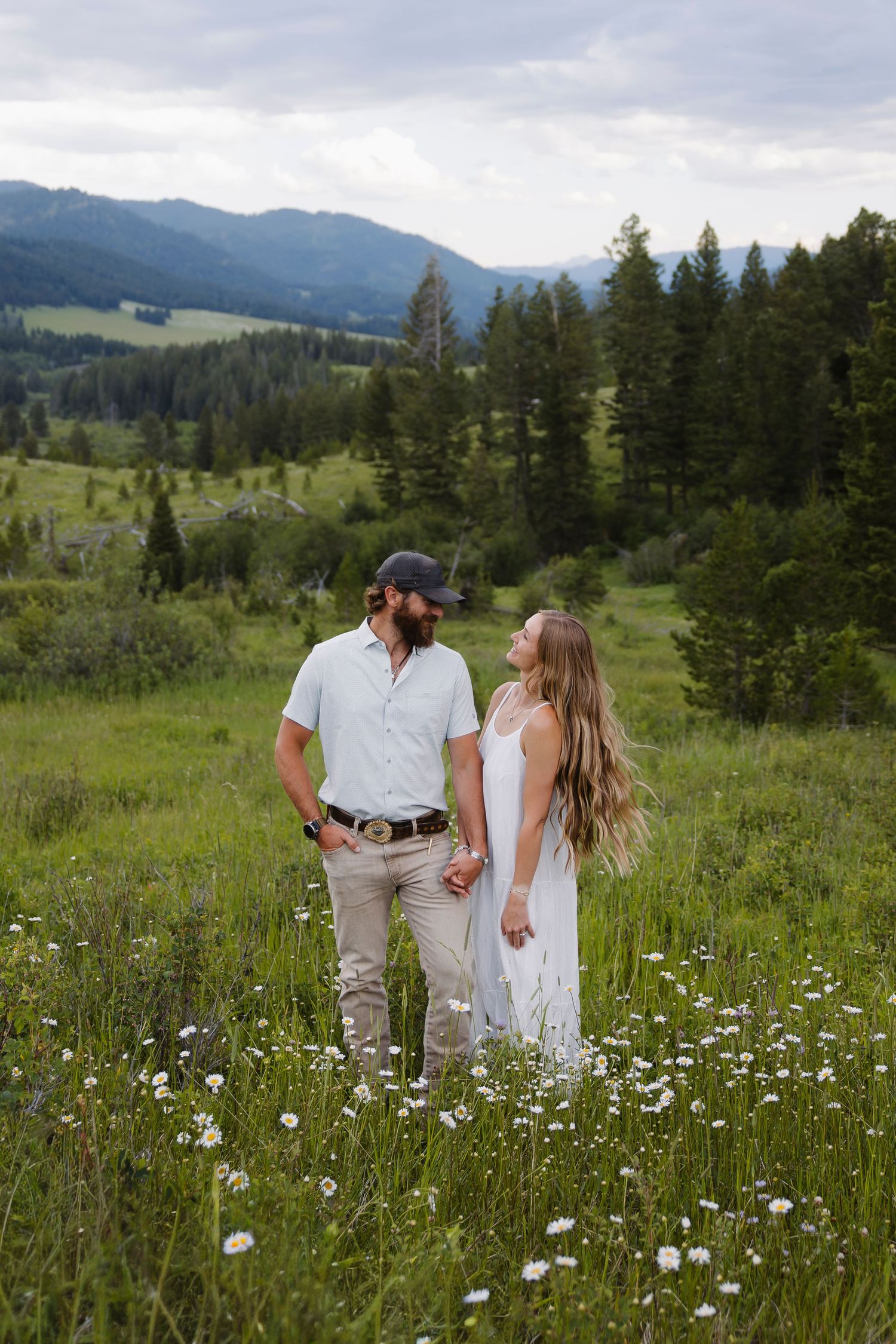 Couple holding hands in a wildflower meadow with mountain forest backdrop, woman in white dress, man in white shirt.