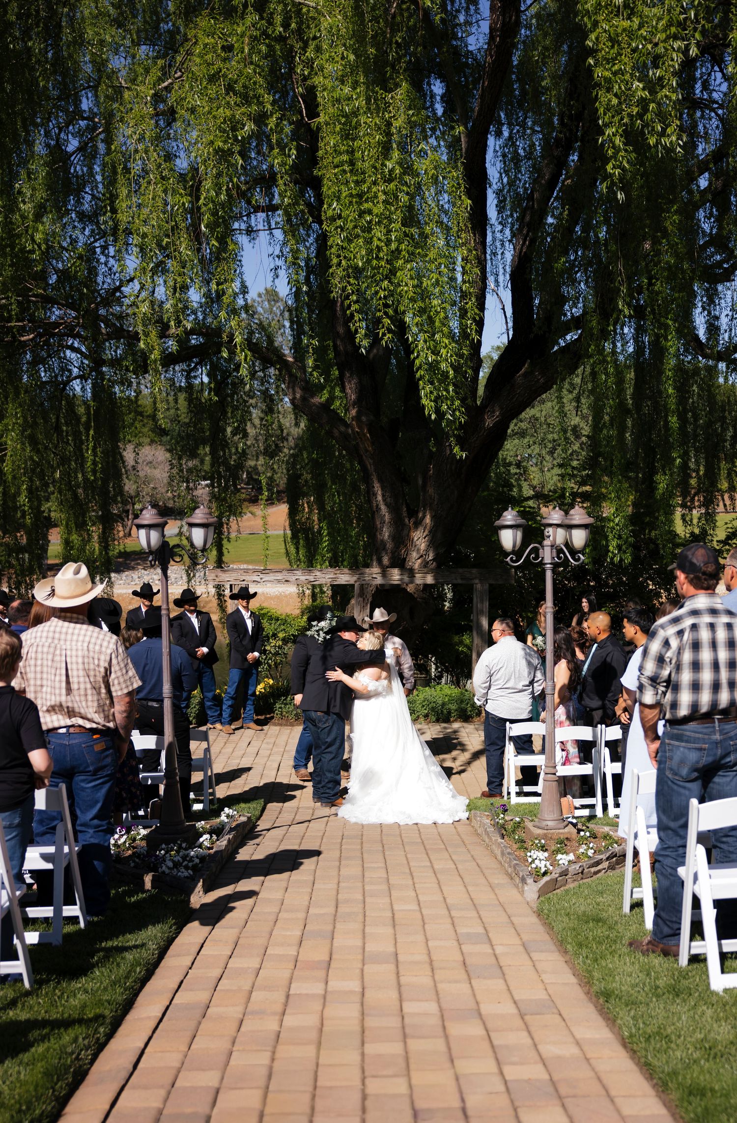 Western wedding ceremony under a large willow tree with guests seated on white chairs along a brick pathway.