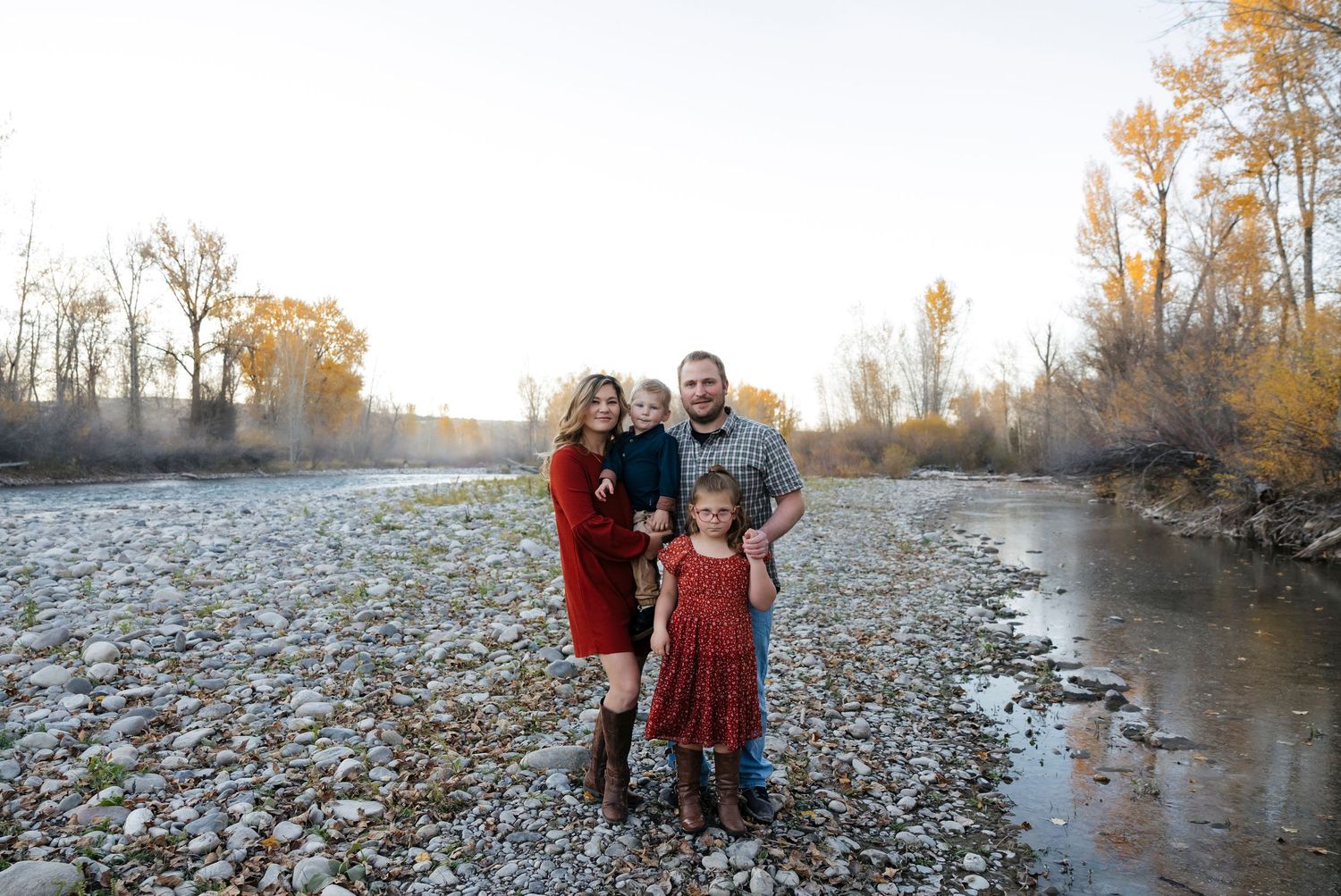 Family of four posing by a rocky riverbank surrounded by autumn trees in golden hour light.