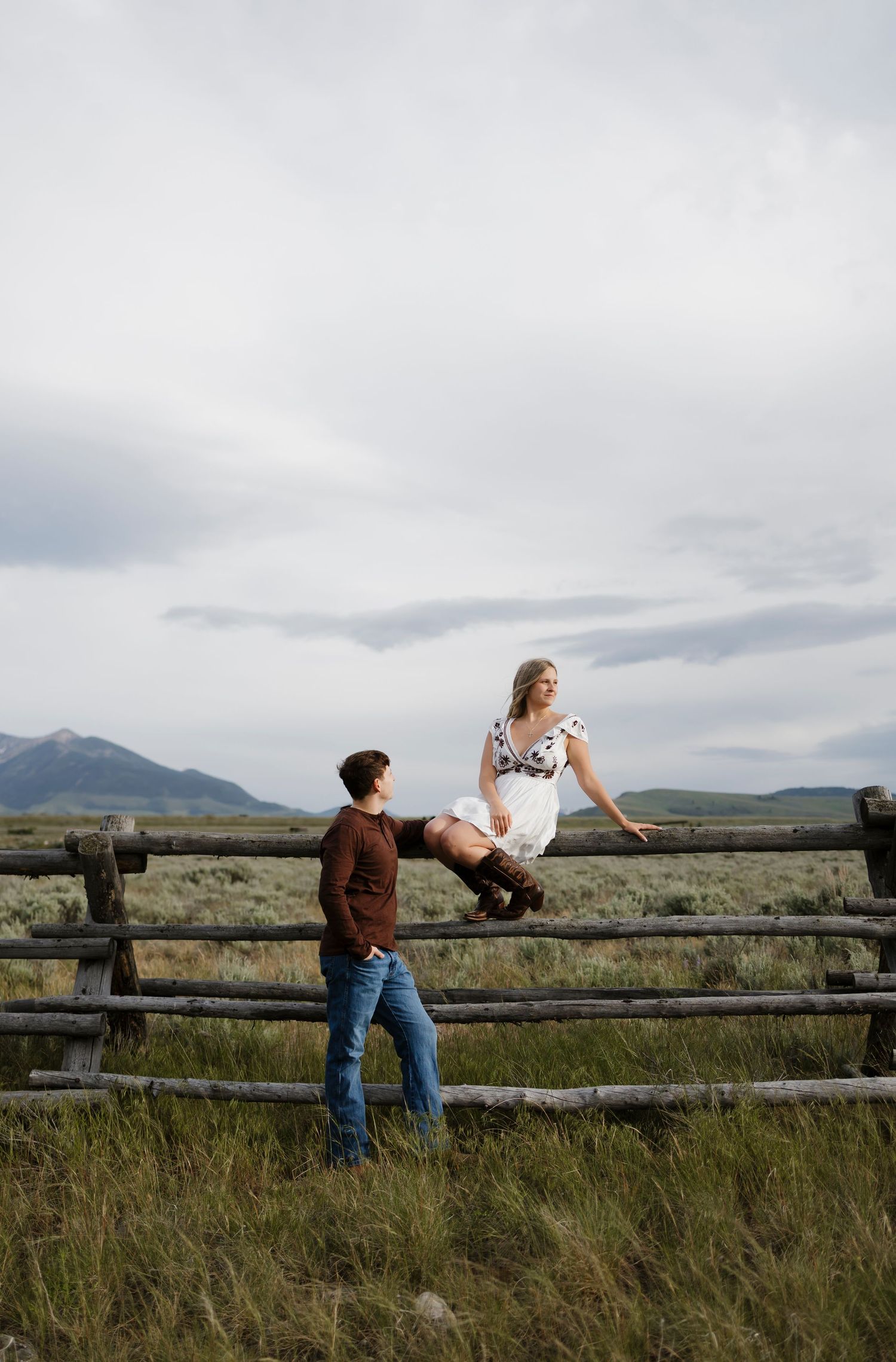 A couple poses at a rustic wooden fence in a mountain meadow, woman sitting on fence in floral dress, man standing below.
