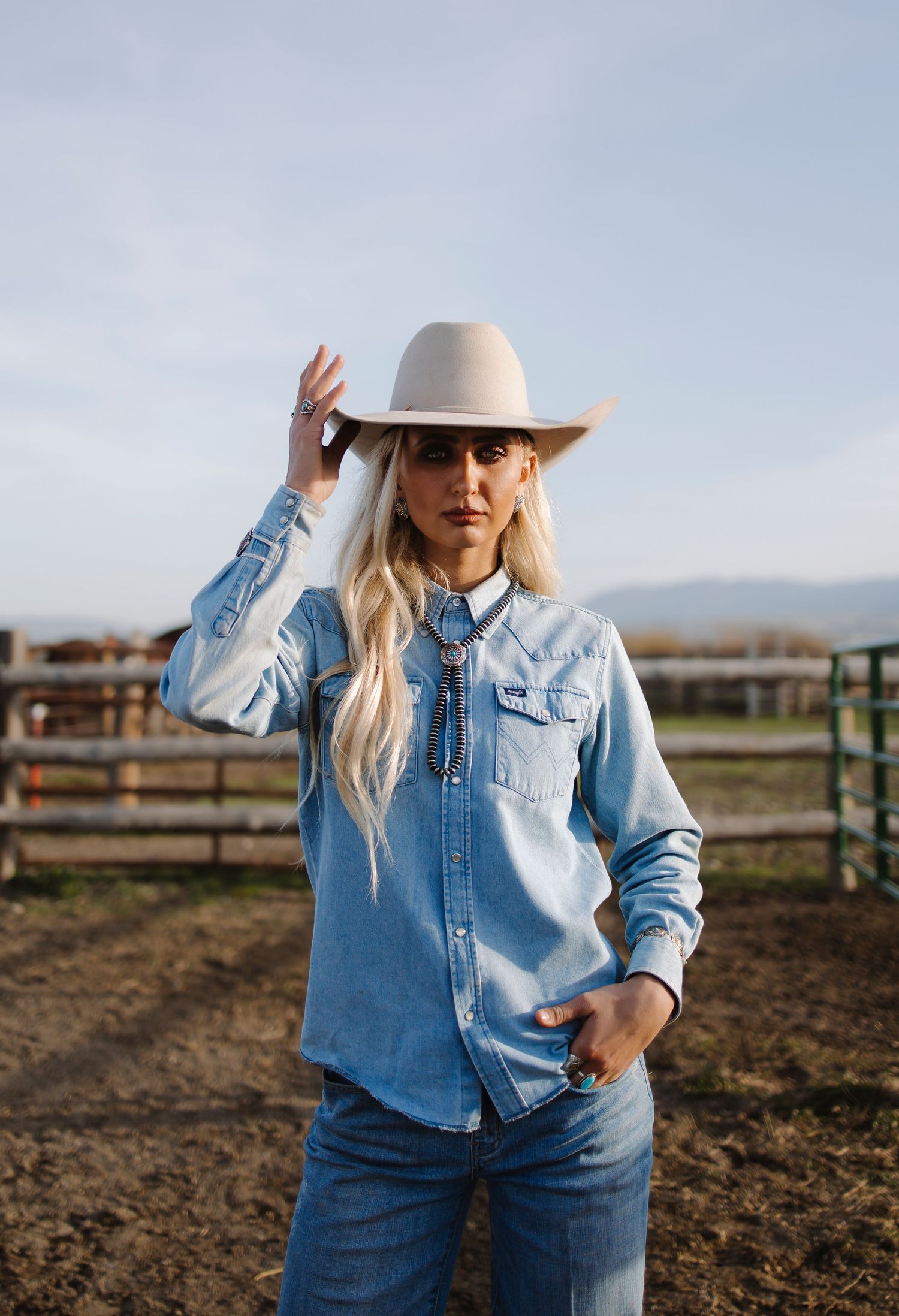 Blonde woman in light denim shirt and beige cowboy hat posing confidently at a ranch.